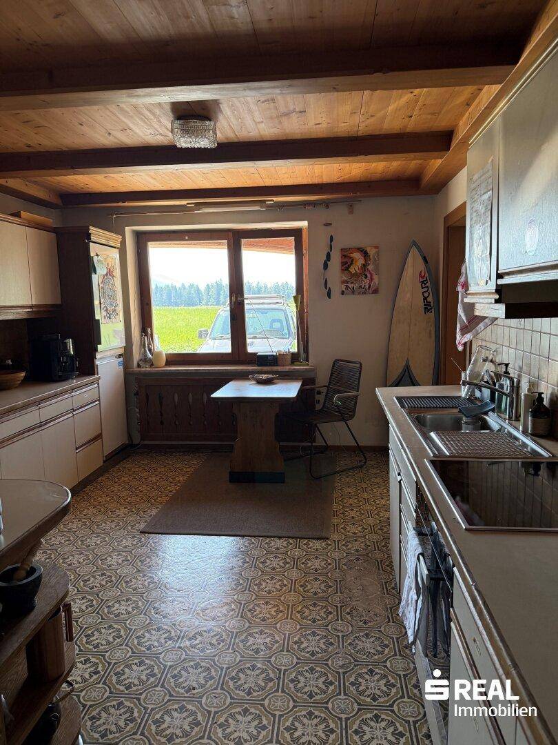 Rustikale Küche mit Holzbalkendecke, gemusterten Fliesen und einem Essbereich am Fenster mit Blick ins Grüne.