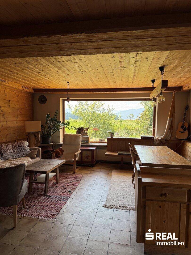 Rustikaler Wohn-Essbereich mit Holzbalkendecke, Fliesenboden und großem Fenster mit weitem Blick ins Grüne.