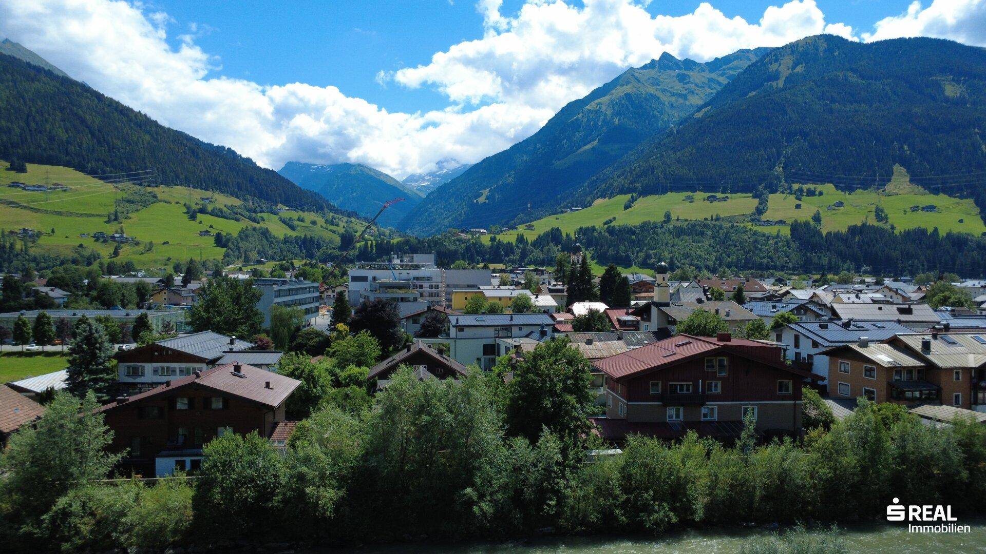 Panoramablick auf eine Stadt im Tal, umgeben von grünen Bergen unter blauem Himmel.