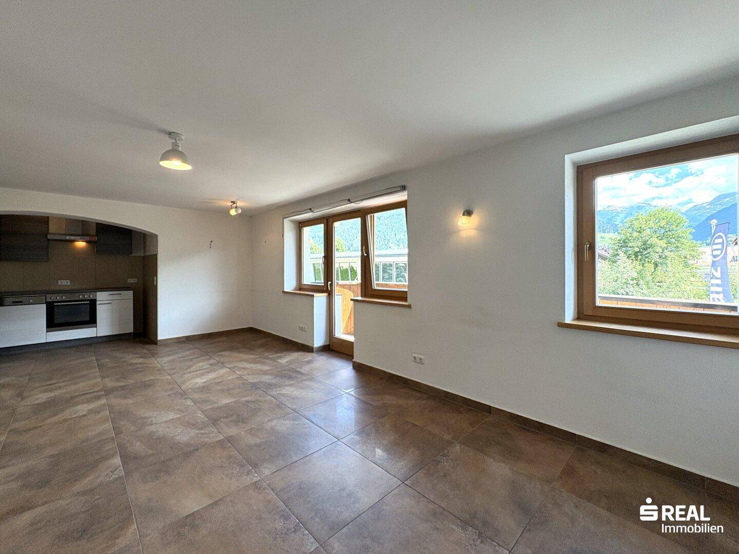 Heller Wohnbereich mit integrierter Küchenzeile, Fliesenboden und Zugang zum Balkon.