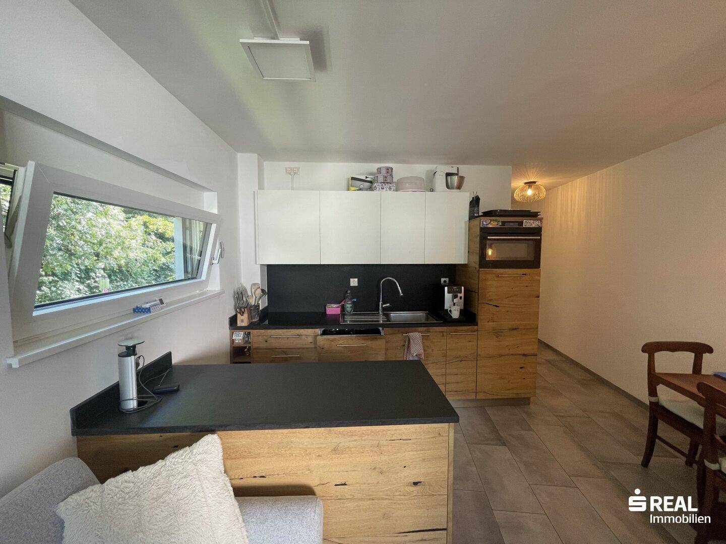 Moderne Küche mit Holzfronten, dunkler Arbeitsplatte und Fenster mit Blick ins Grüne.