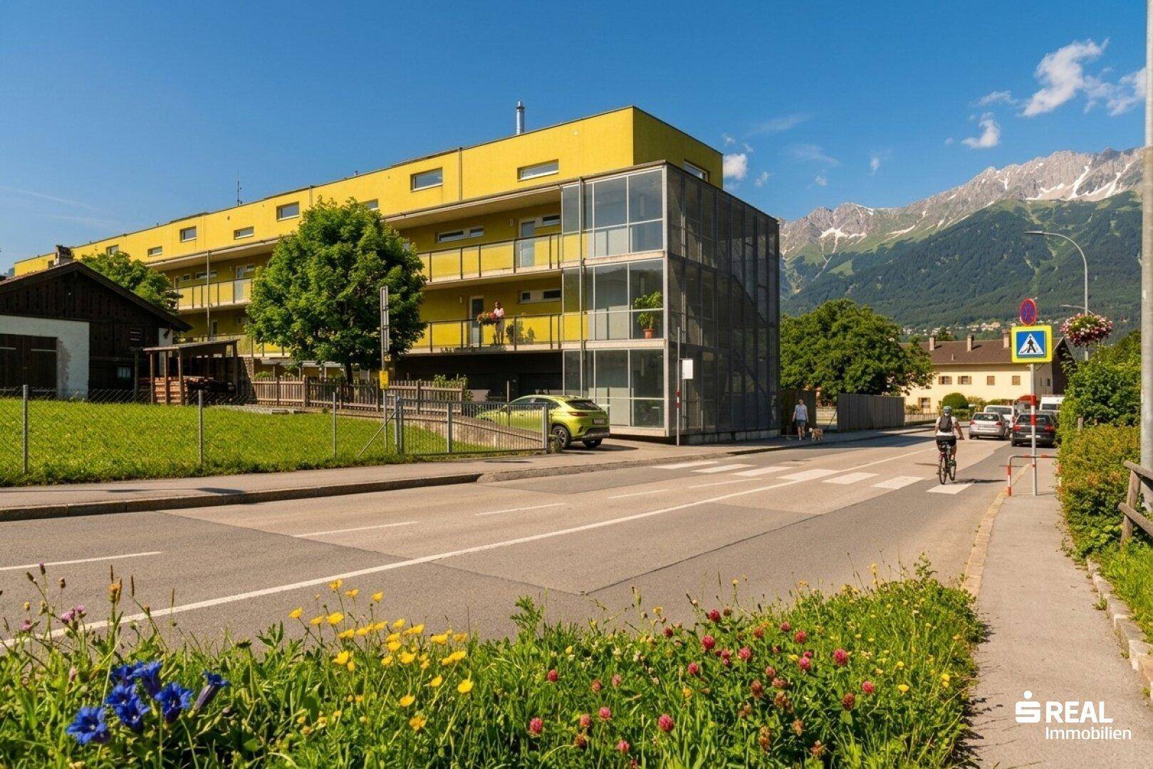 Modernes gelbes Gebäude mit Glasfassade und Balkonen, umgeben von Grünflächen und mit Bergblick.