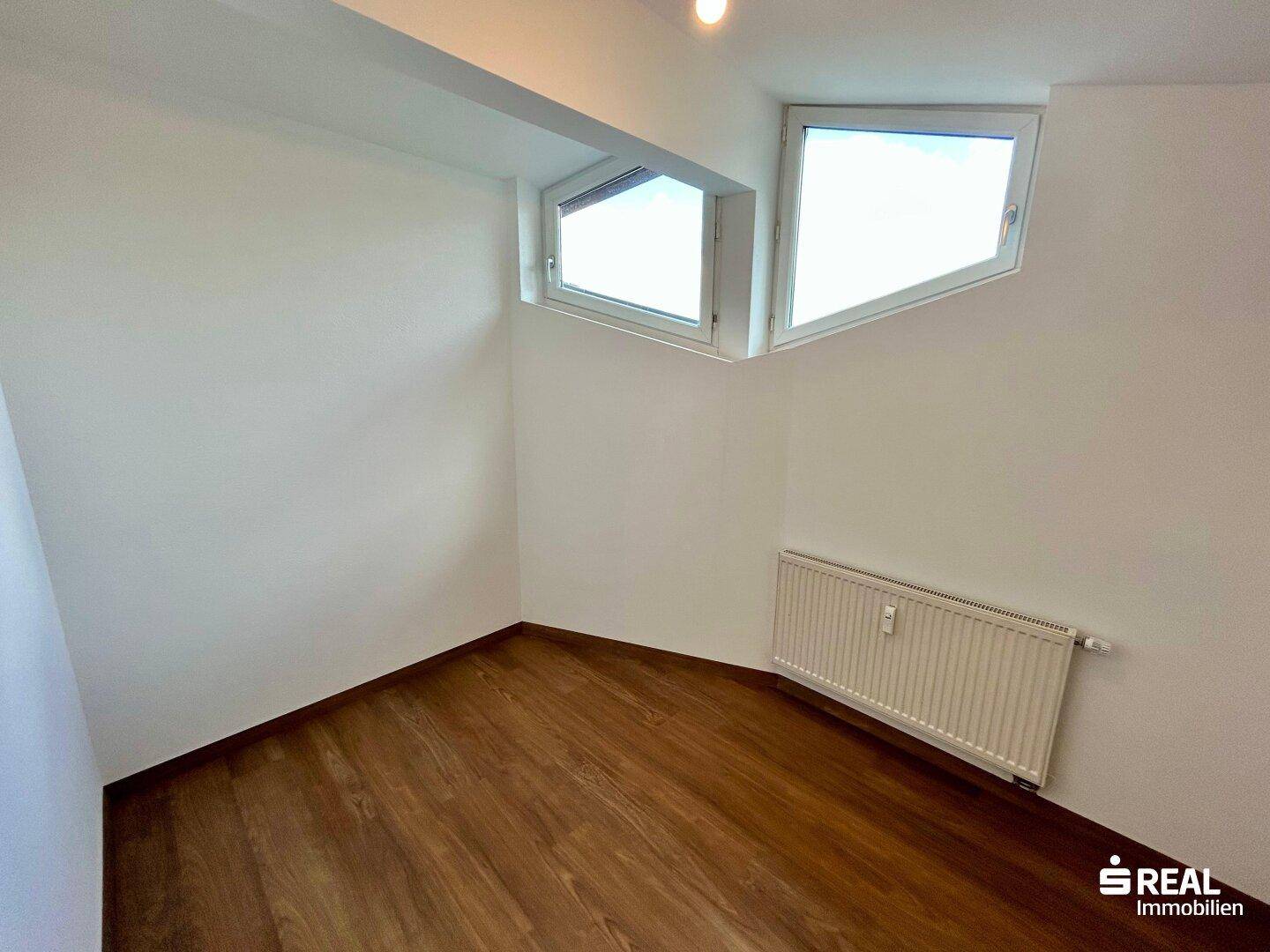 Helles Schlafzimmer mit Laminatboden und zwei schrägen Fenstern für natürlichen Lichteinfall.