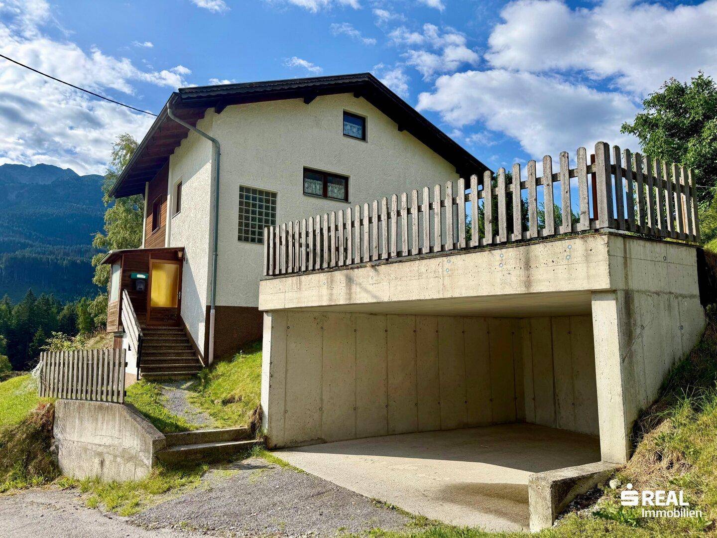 Haus mit integrierter Garage und Holzgeländer, umgeben von grüner Landschaft.