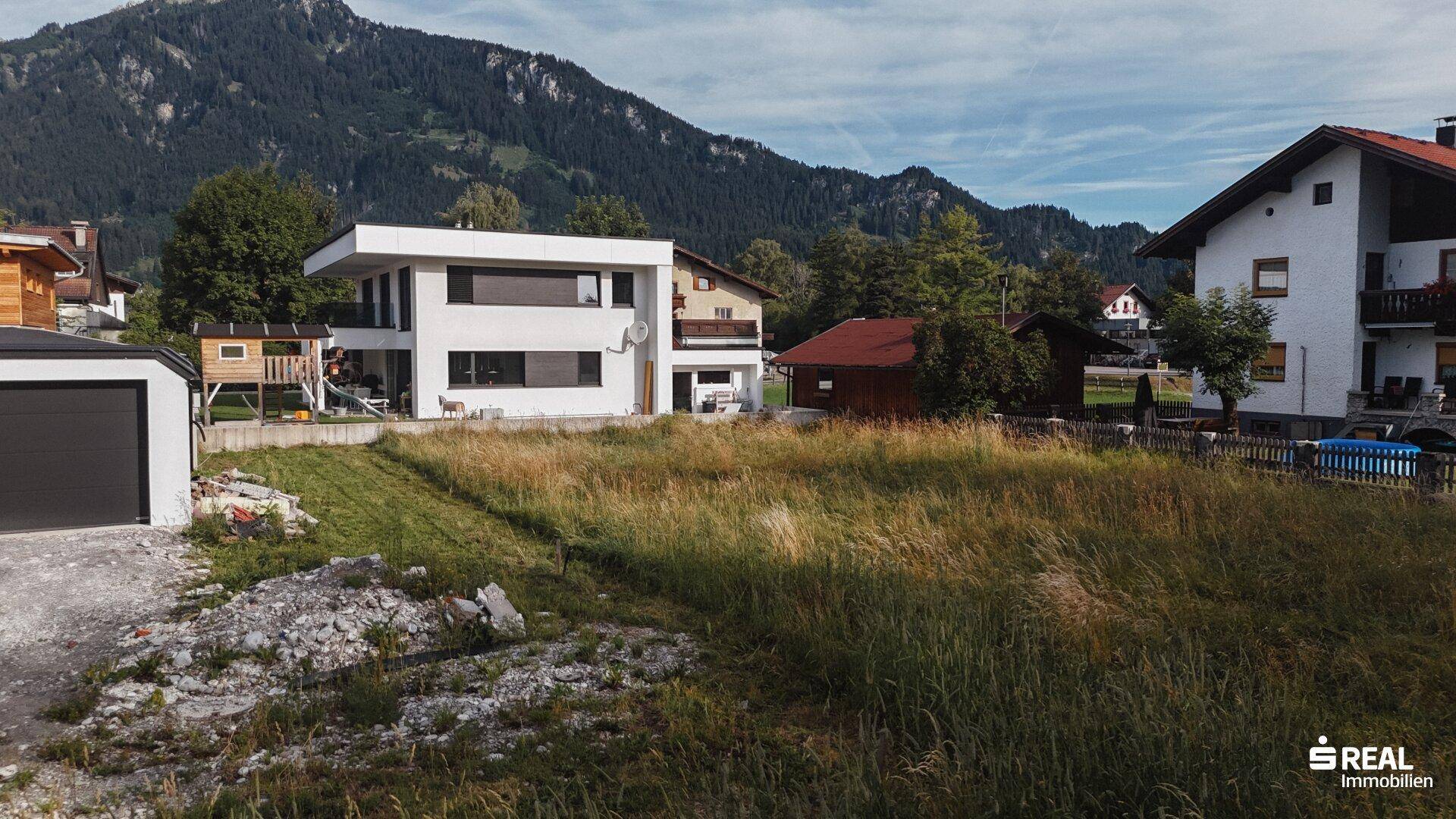 Blick über ein unbebautes Grundstück mit hohen Gräsern und Bergen im Hintergrund.