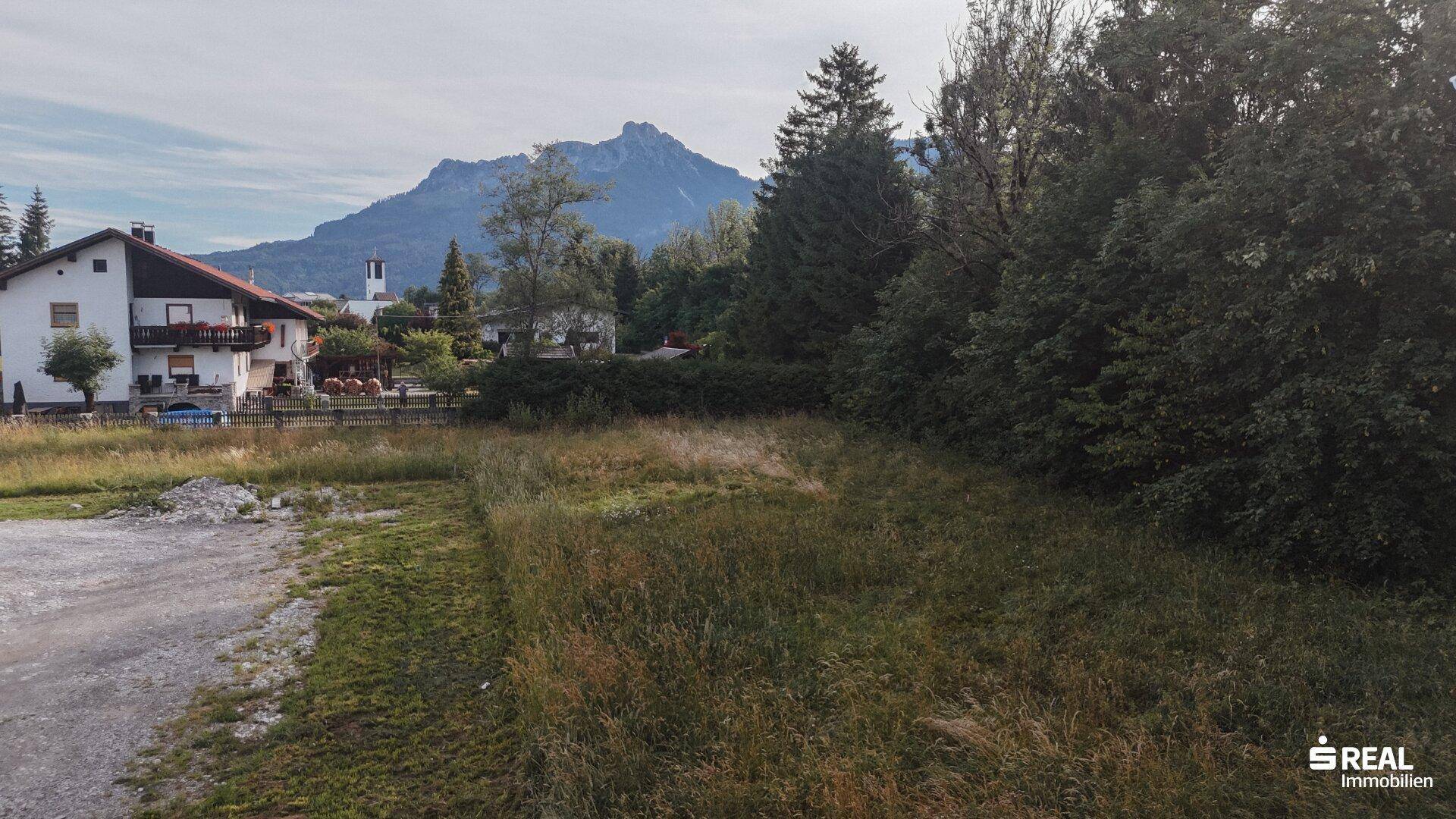 Unbebautes Grundstück mit Gräsern, umgeben von Häusern und einer beeindruckenden Bergkulisse.