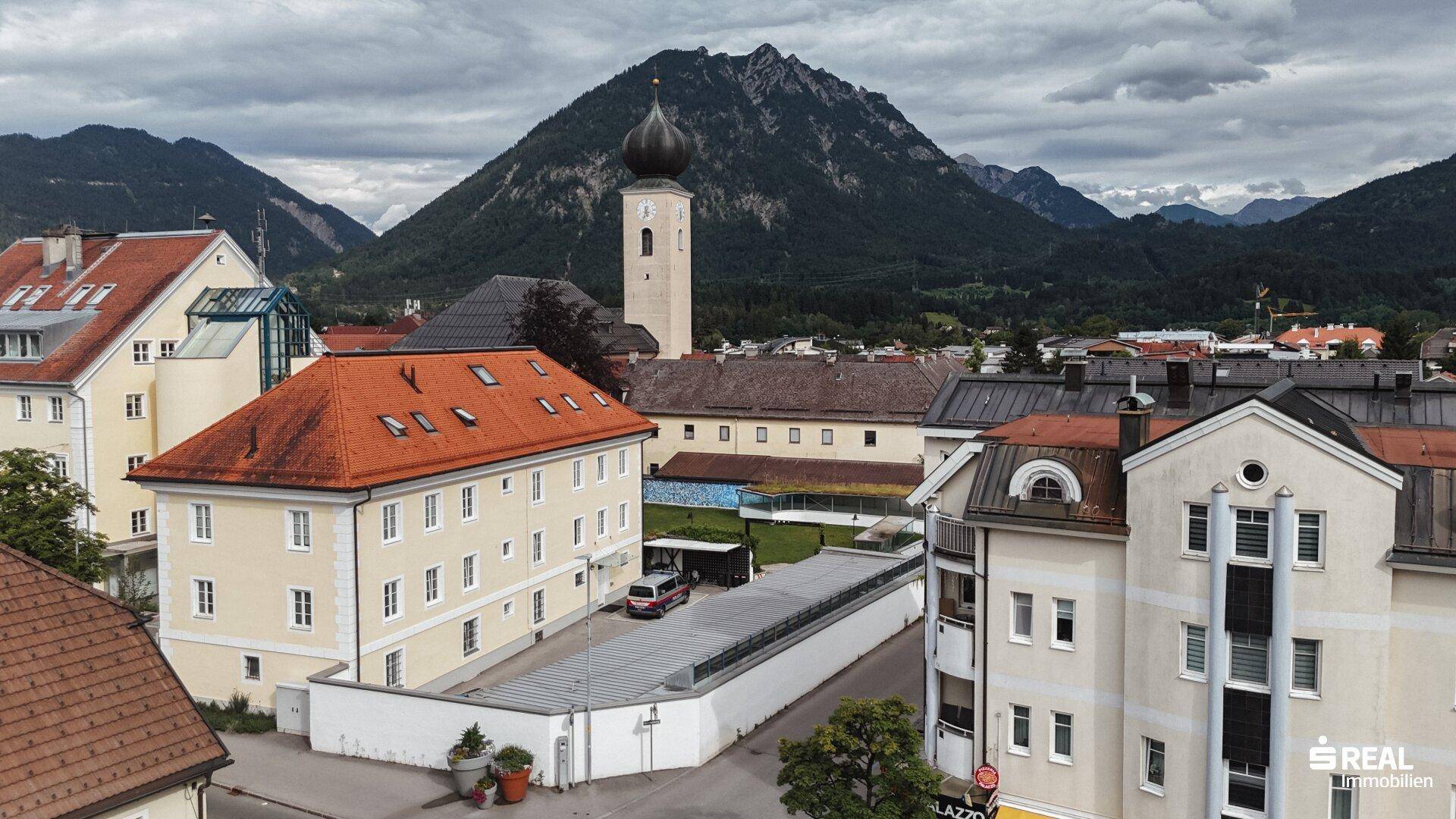 Luftaufnahme einer Stadt mit Kirchturm und traditionellen Gebäuden, umgeben von majestätischen Bergen.