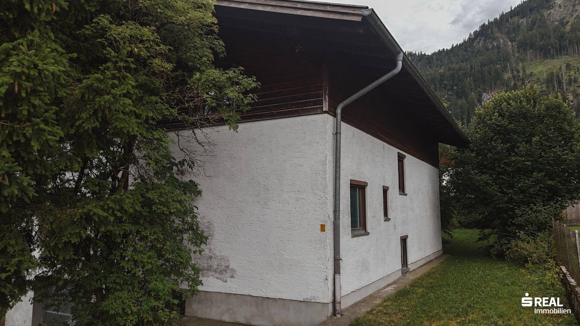 Rückansicht eines Hauses mit weißer Fassade und schmalen Fenstern, umgeben von üppigem Grün.