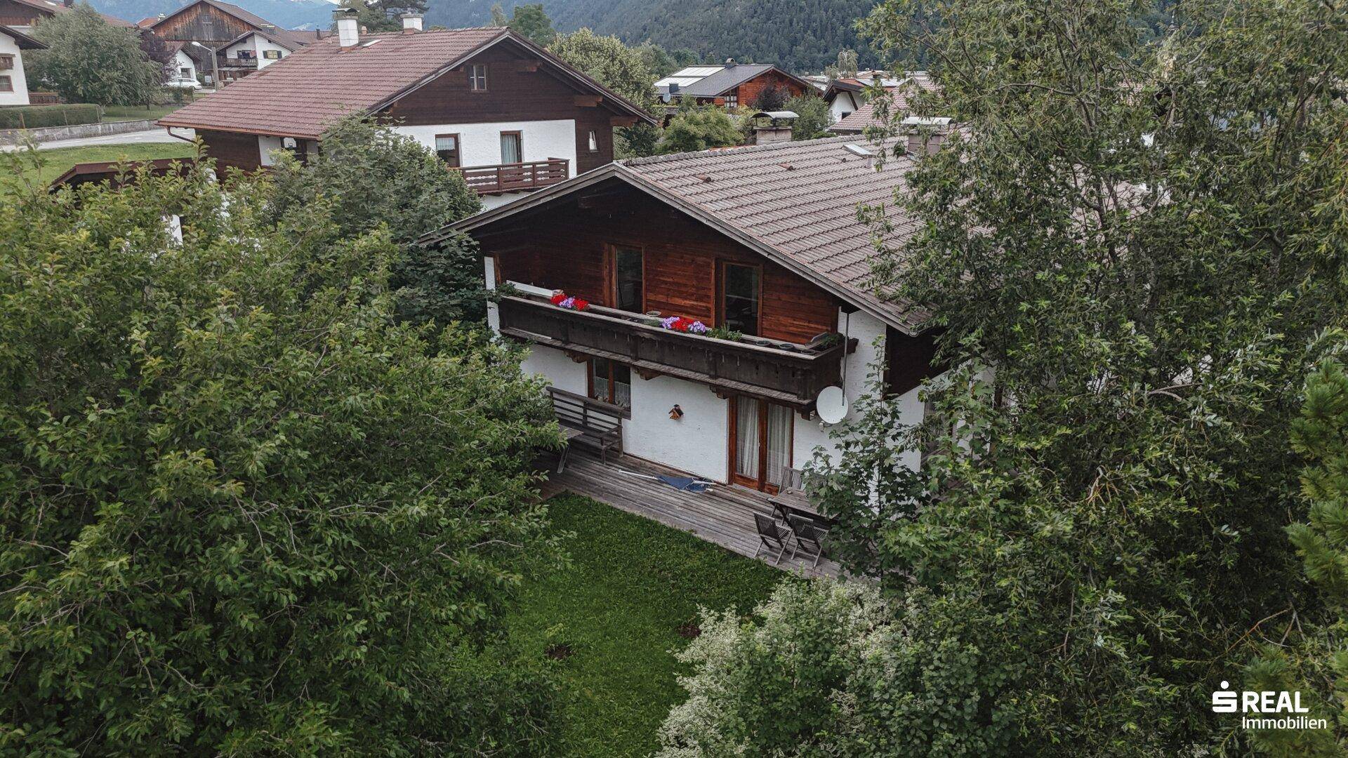 Vogelperspektive eines Wohnhauses mit Holzbalkon und Garten, umgeben von hohen Bäumen.