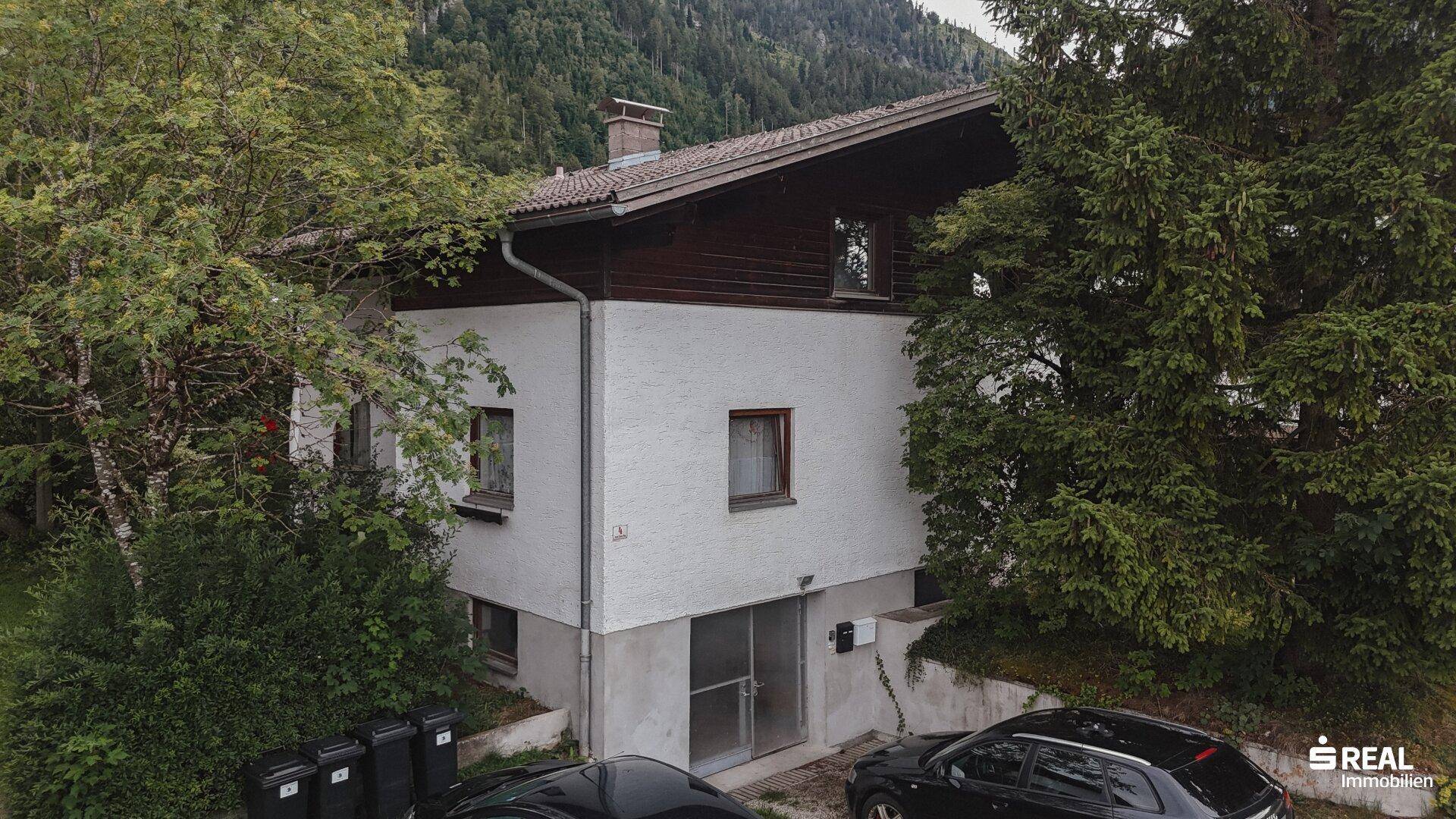 Seitenansicht eines Wohnhauses mit weißer Fassade und kleinen Fenstern, umgeben von Bäumen.