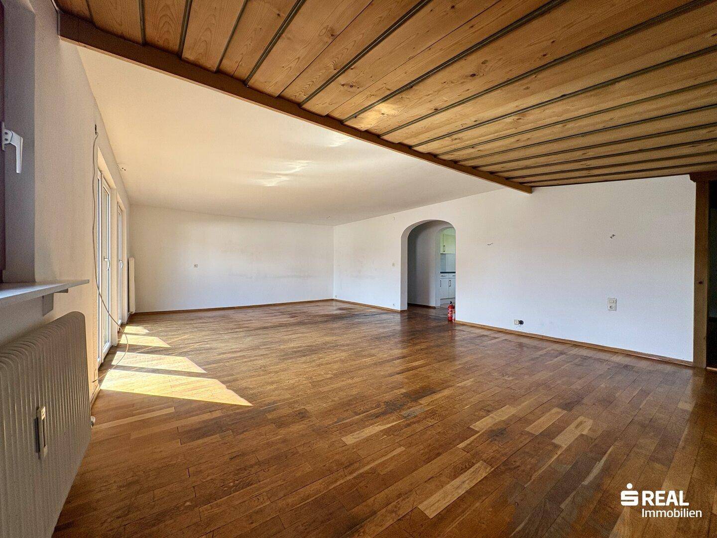 Geräumiges Wohnzimmer mit Holzbalkendecke, Parkettboden und einem Durchgang zu einem weiteren Raum.