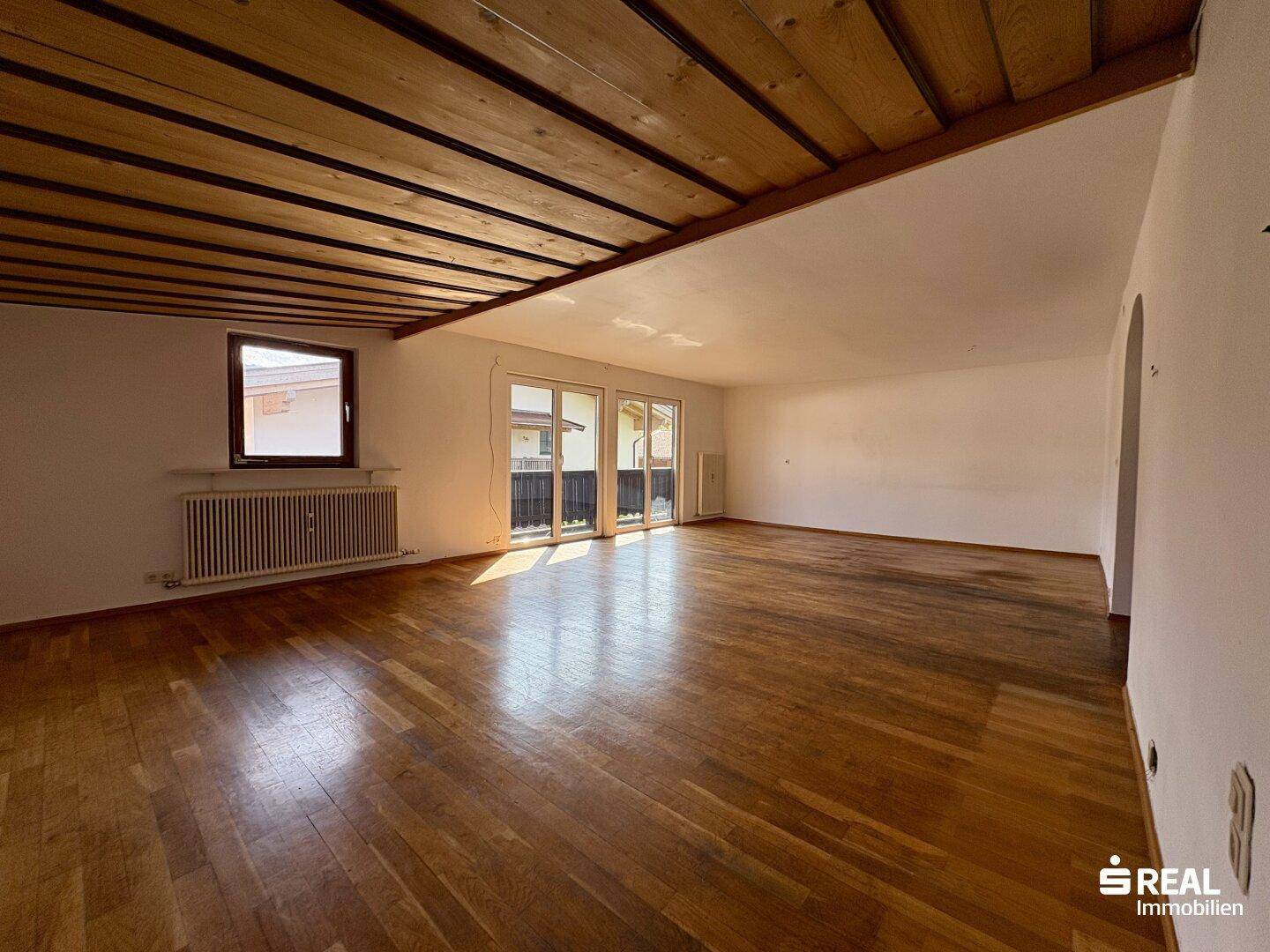 Geräumiges, unmöbliertes Wohnzimmer mit Holzbalkendecke, Parkettboden und Zugang zum Balkon.