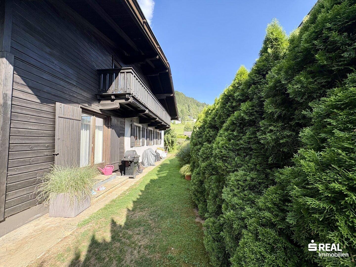 Seitenansicht des Hauses mit schmalem Weg, hoher Hecke und Holzbalkon im Obergeschoss.