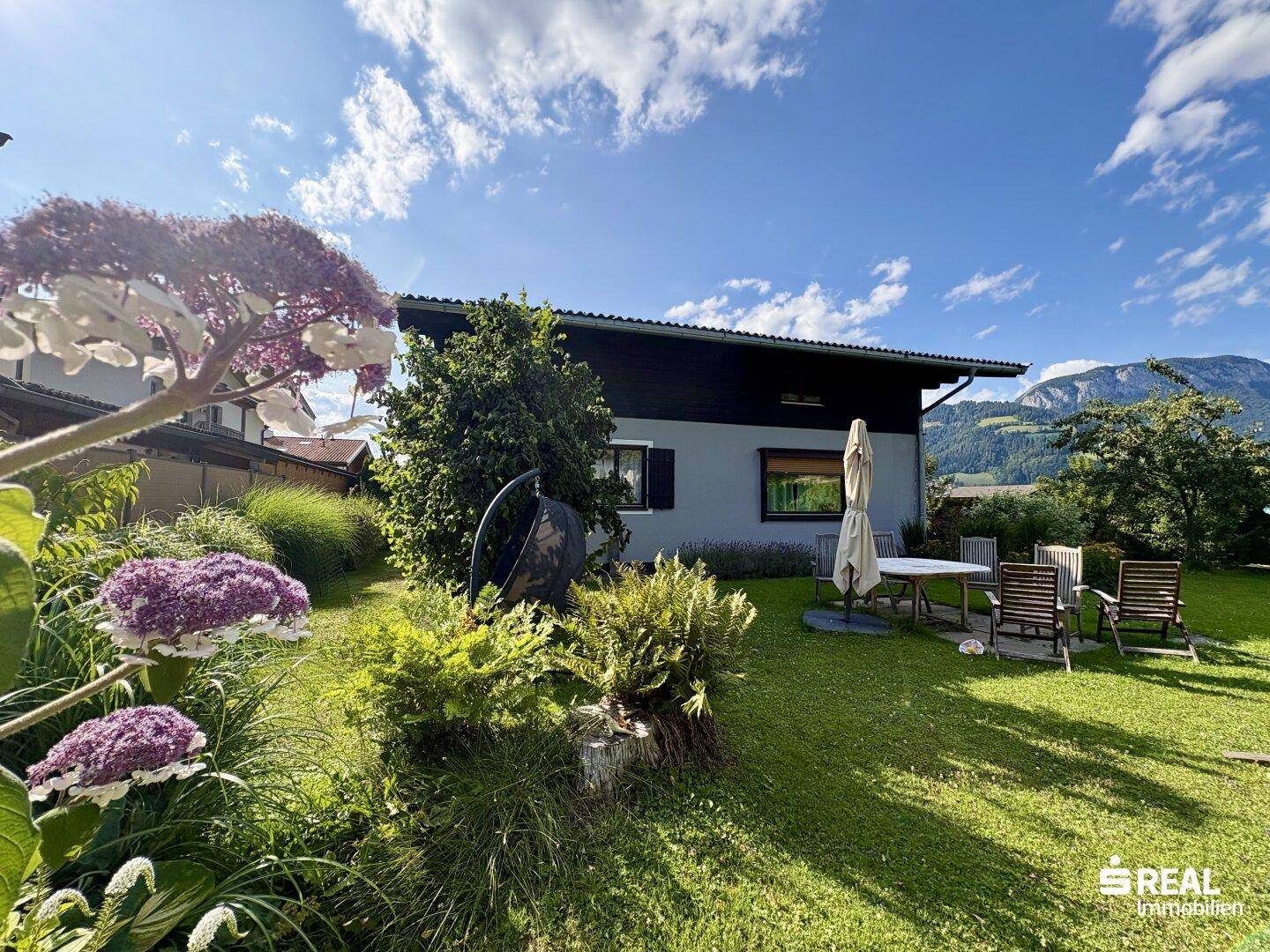 Sonniger Gartenbereich mit Sitzgelegenheiten, Blumenbeeten und Blick auf die Berge.