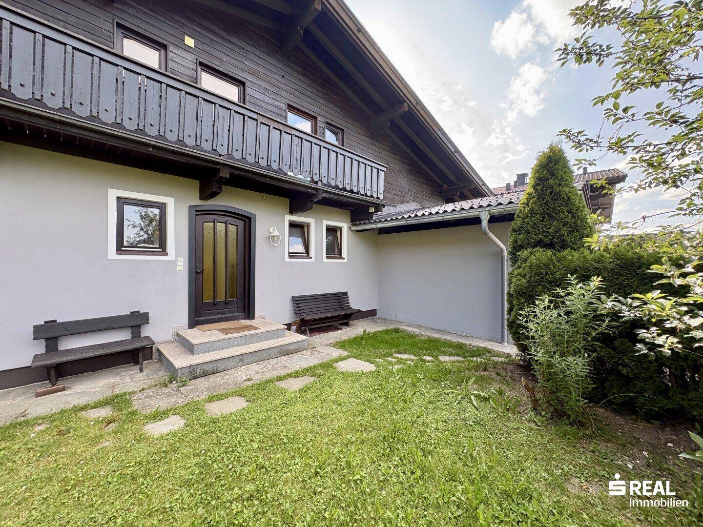Eingangsbereich des Hauses mit kleiner Grünfläche, Steinplatten und Holzbalkon im Obergeschoss.