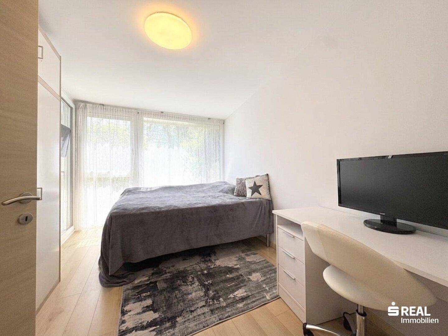 Gemütliches Schlafzimmer mit Holzboden, großem Fenster und modernem Schreibtisch.
