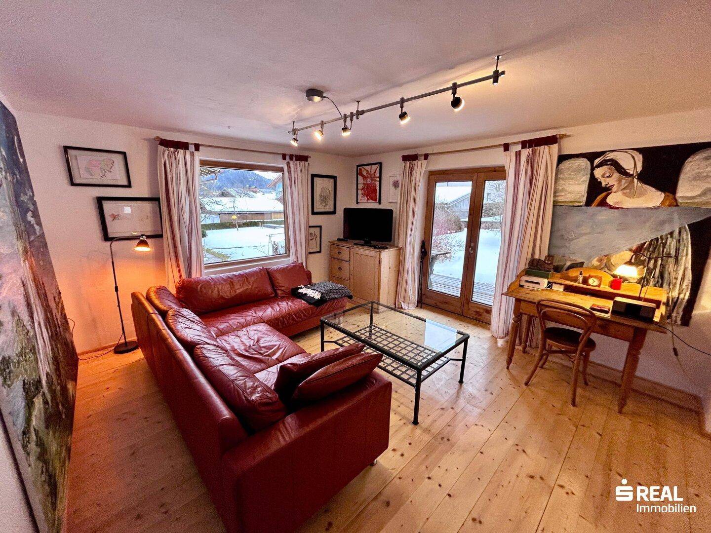 Gemütliches Wohnzimmer mit rotem Ledersofa, Holzboden und Zugang zum Balkon.