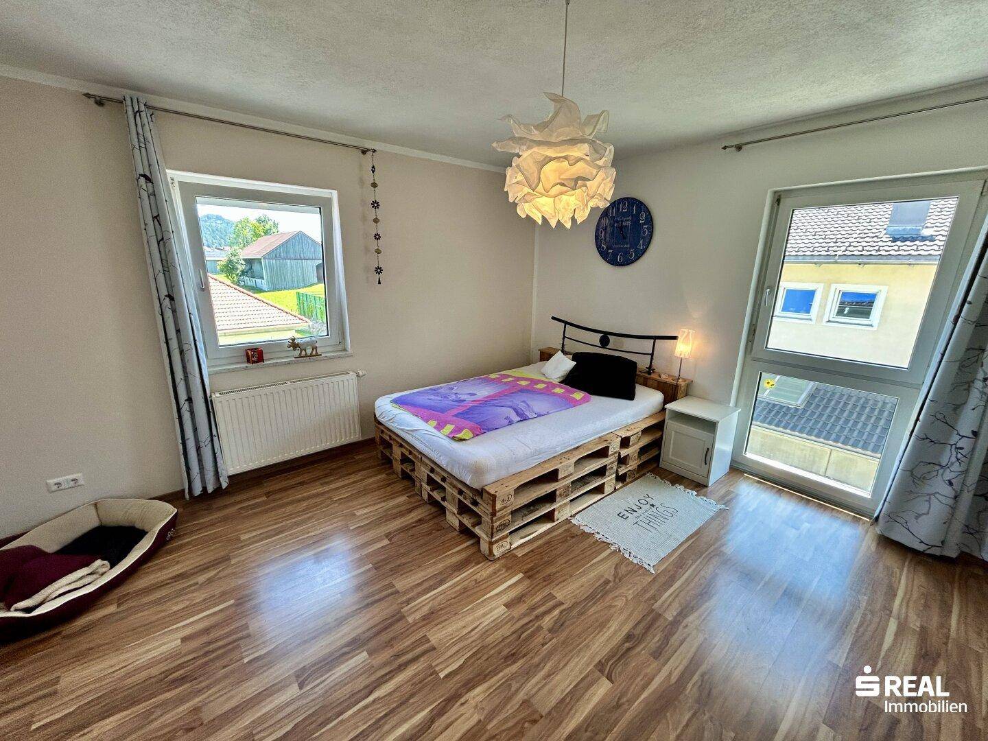 Schlafzimmer mit kreativem Palettenbett, Holzboden und großem Fenster für Helligkeit.