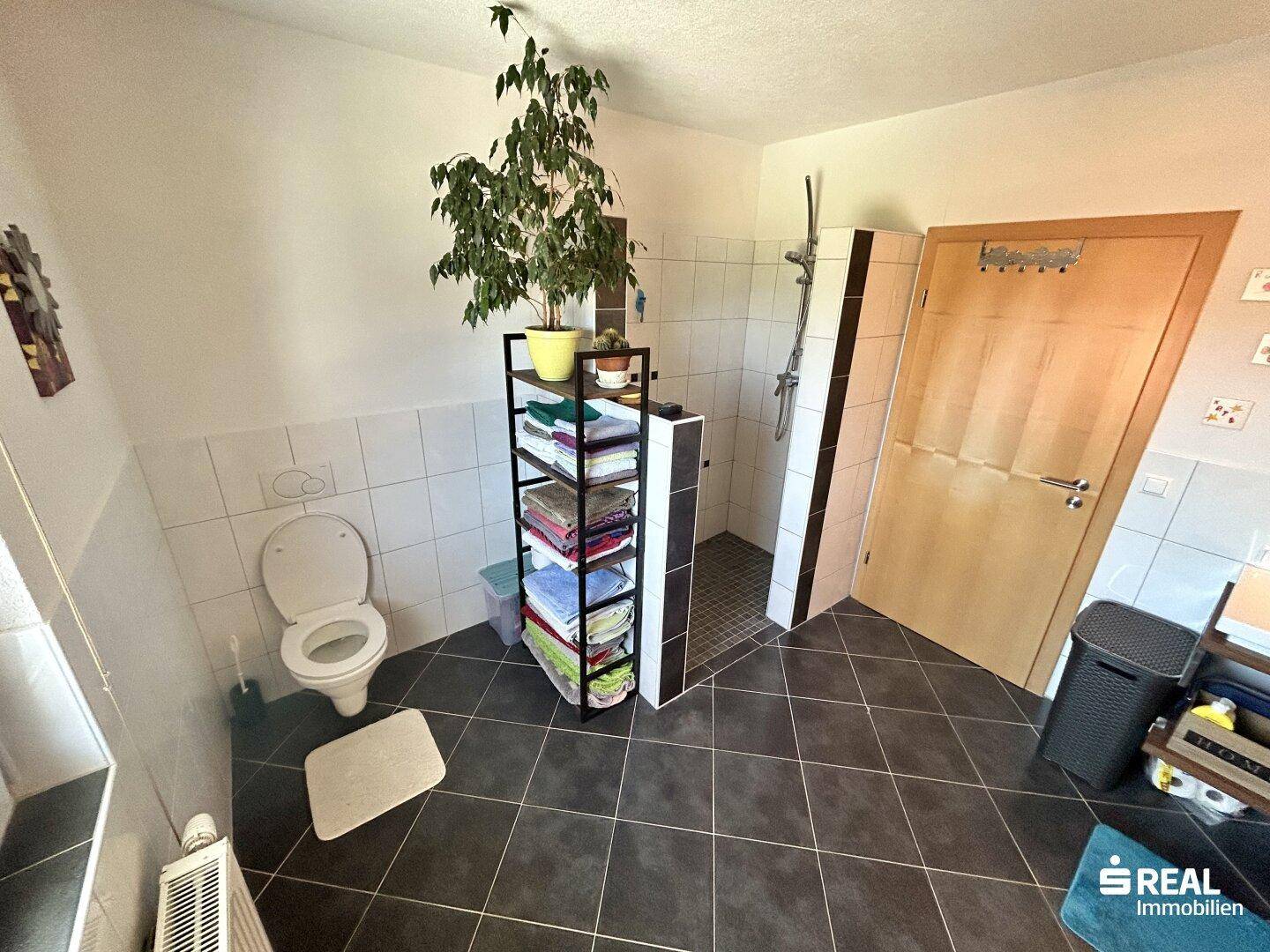 Geräumiges Badezimmer mit großer Dusche, WC und dunklen Fliesen.