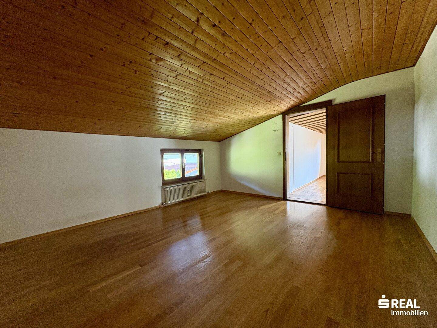 Gemütliches Dachzimmer mit Holzboden, schräger Decke, Fenster und Heizkörper unter dem Fenster.