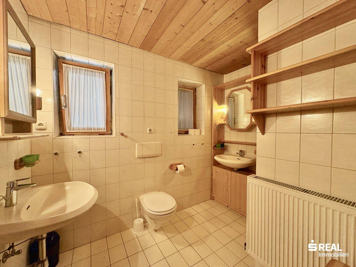 Funktionales Badezimmer mit hellen Fliesen, Holzdecke, zwei Waschbecken und einem Fenster für Tageslicht.