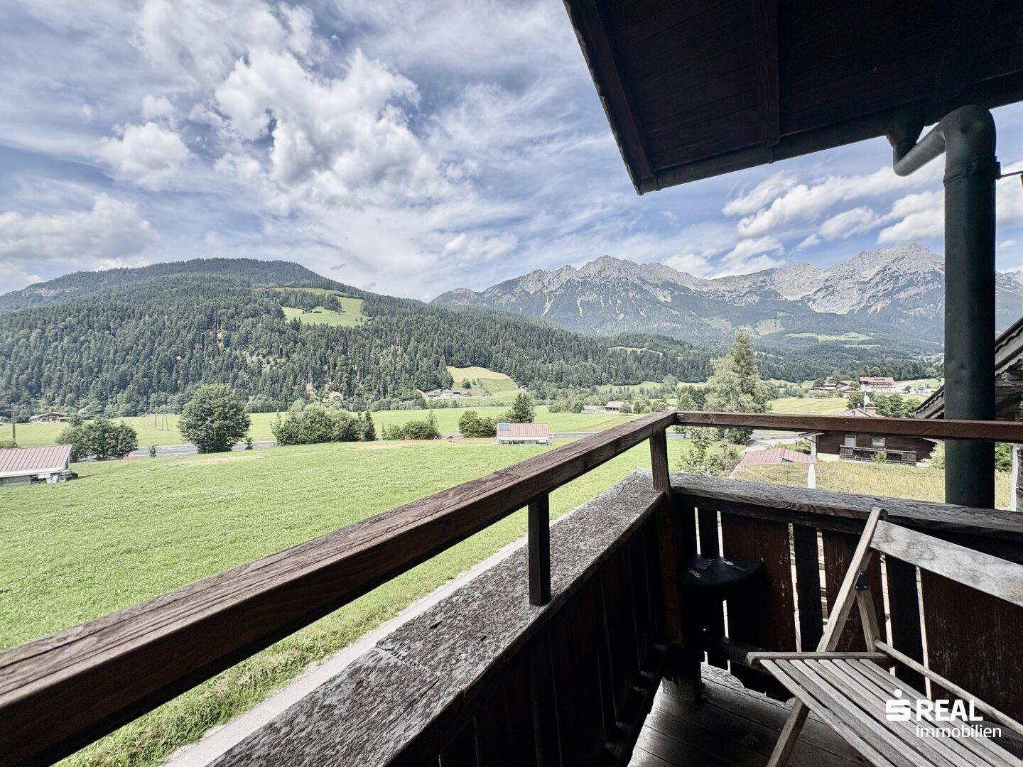 Breiter Holzbalkon mit weitem Blick über grüne Wiesen und die beeindruckende Bergkulisse.
