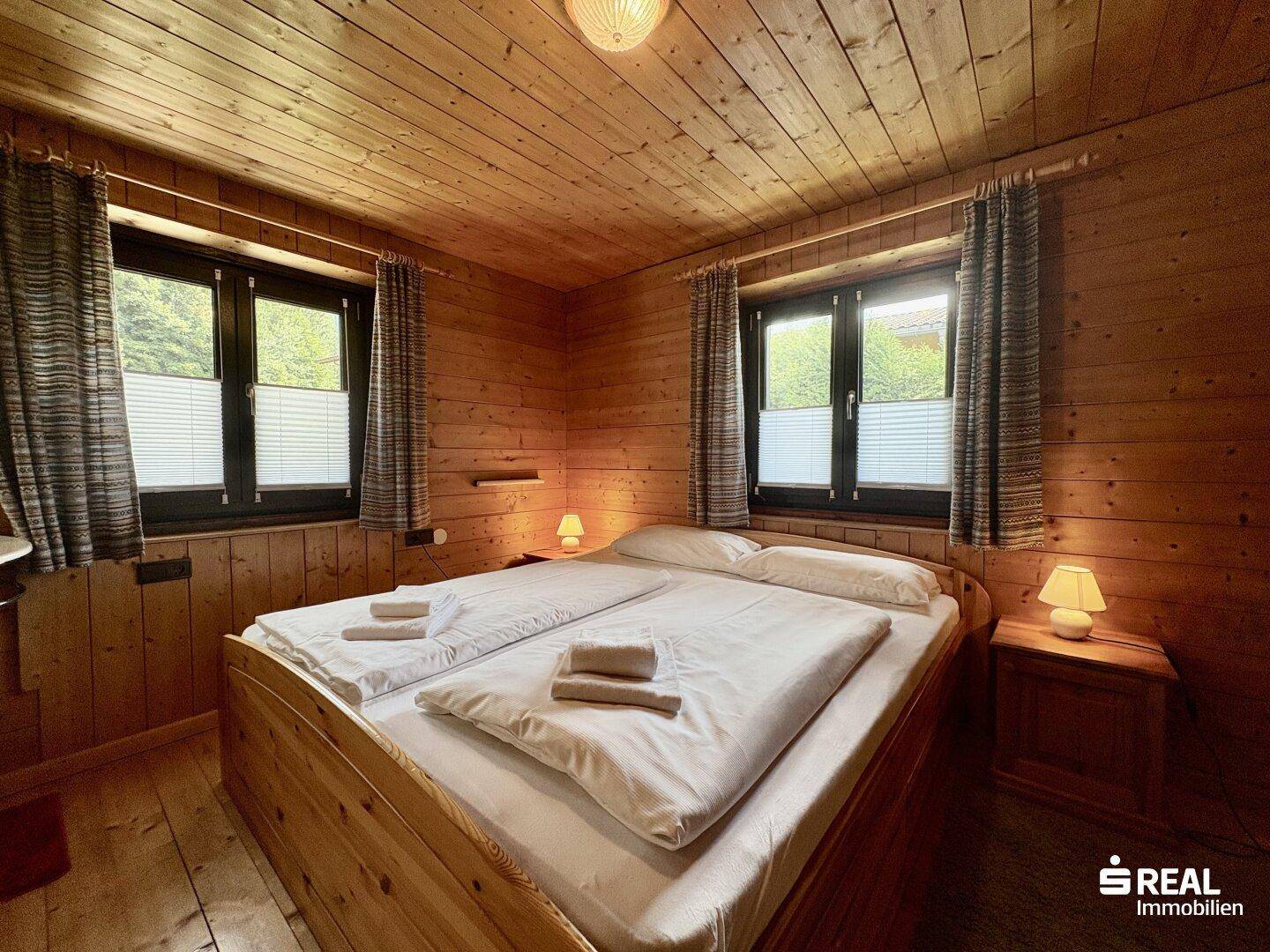 Helles Schlafzimmer mit Holztäfelung, Doppelbett und zwei Fenstern, die viel Tageslicht hereinlassen.