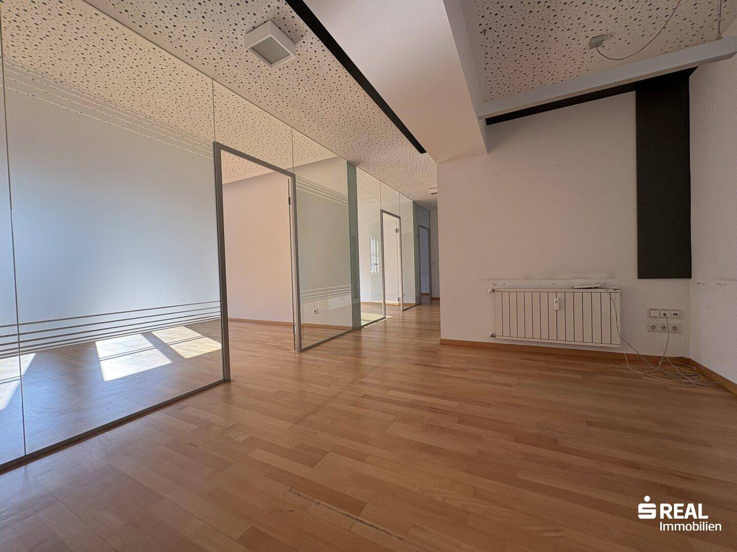 Geräumiges, leeres Büro mit Glaswänden, Holzboden und einer Akustikdecke, gut beleuchtet.