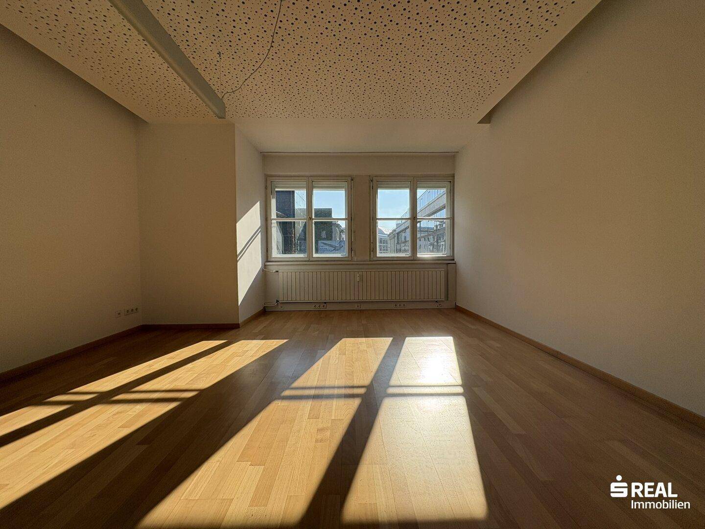 Heller Büroraum mit Holzboden, zwei großen Fenstern und einem Heizkörper, durchflutet von Sonnenlicht.