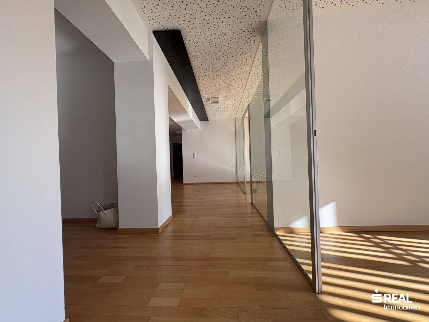 Langer Flur mit Holzboden, Glaswänden und einer Akustikdecke, der zu weiteren Büroräumen führt.
