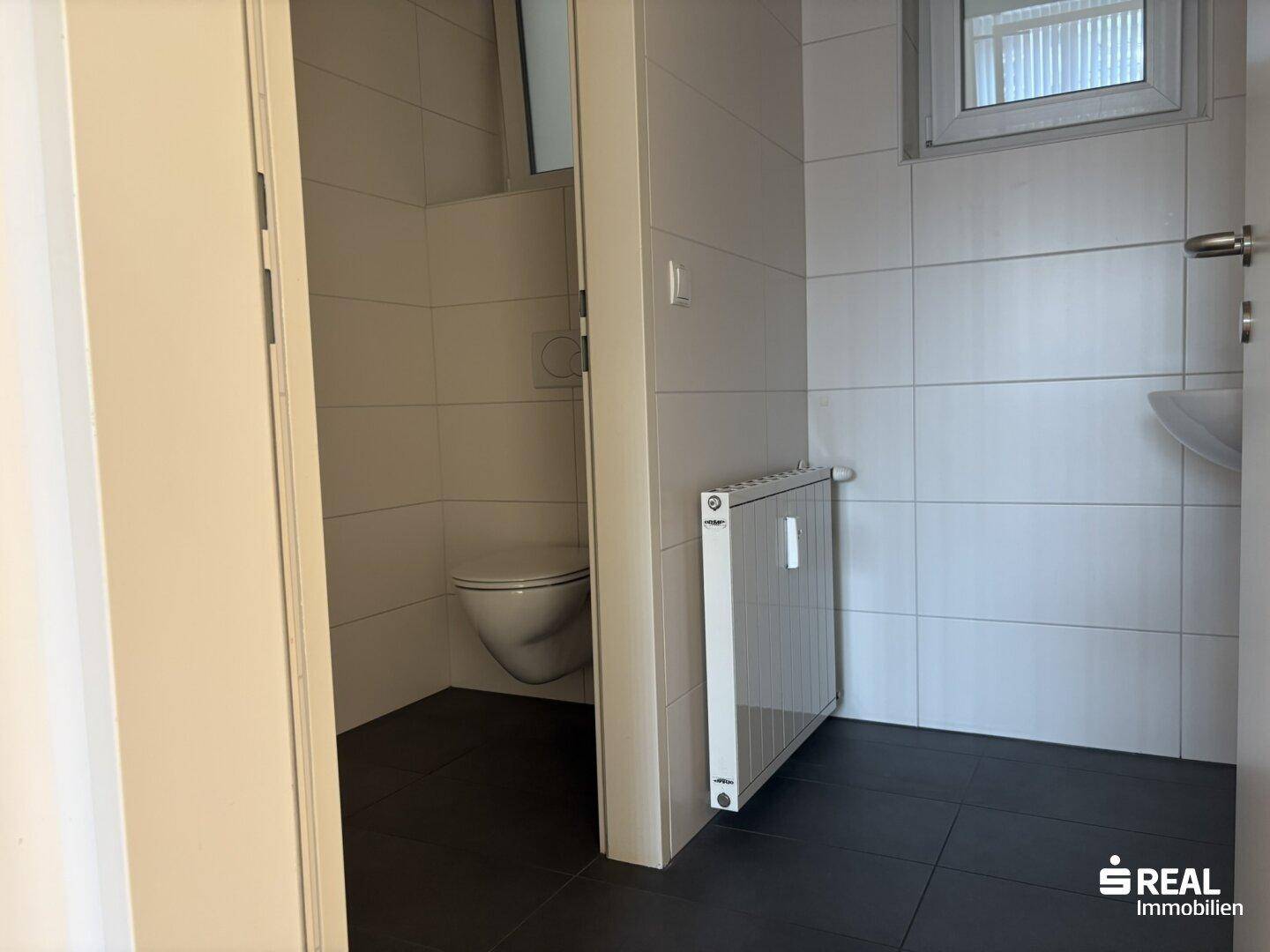 Kleiner, sauberer Toilettenraum mit weißen Wandfliesen, dunklem Boden und einem Fenster für Tageslicht.