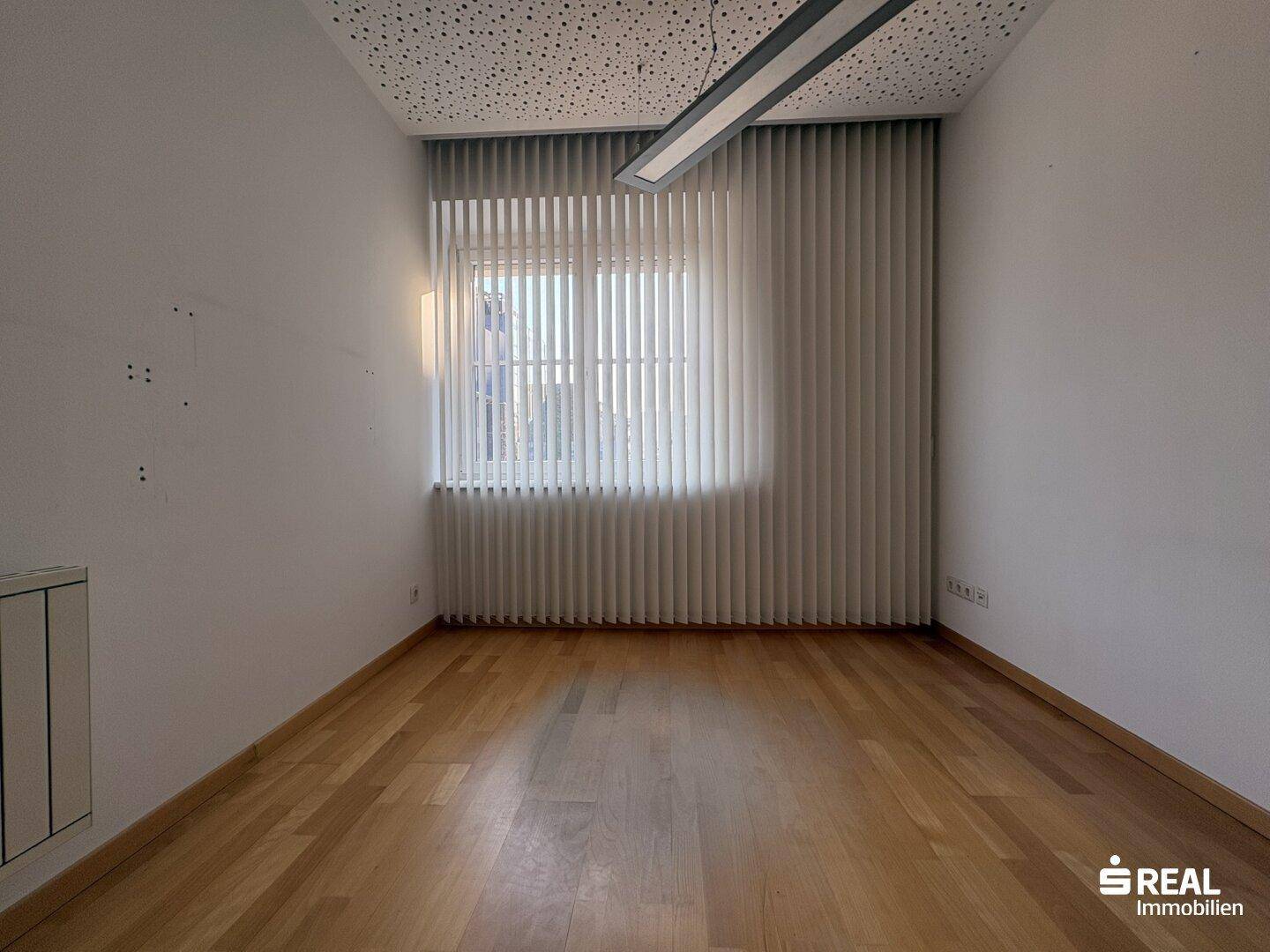 Heller Büroraum mit Holzboden, Fenster mit vertikalen Jalousien und einer Akustikdecke.