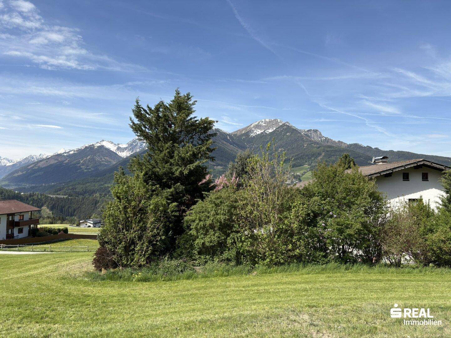 Malerische Landschaft mit grünen Wiesen, Bäumen und schneebedeckten Bergen unter einem strahlend blauen Himmel.