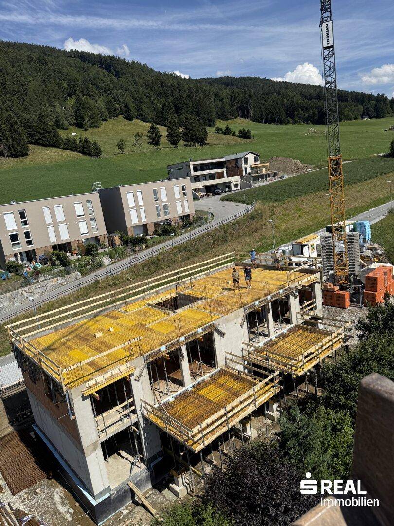 Luftaufnahme eines Neubaus im Bau mit Dachkonstruktion und Gerüst, umgeben von grünen Hügeln und anderen Gebäuden.