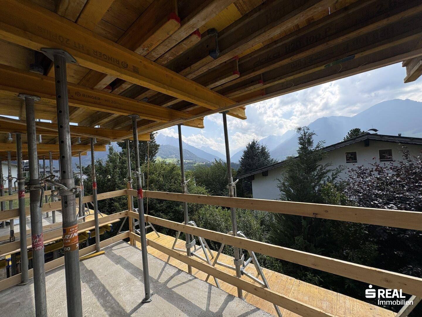 Blick von einem im Bau befindlichen Balkon mit Holzgerüst und Betonboden, umgeben von Bäumen und Bergen.