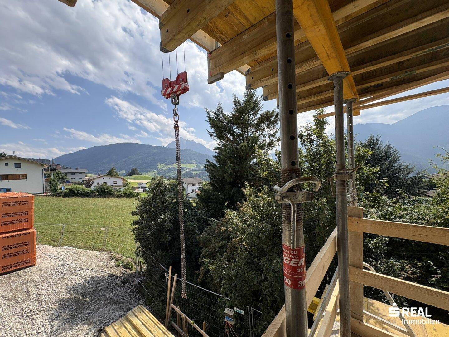 Blick von der Baustelle mit Gerüst und Kranhaken, umgeben von grünen Bäumen und einer beeindruckenden Bergkulisse.
