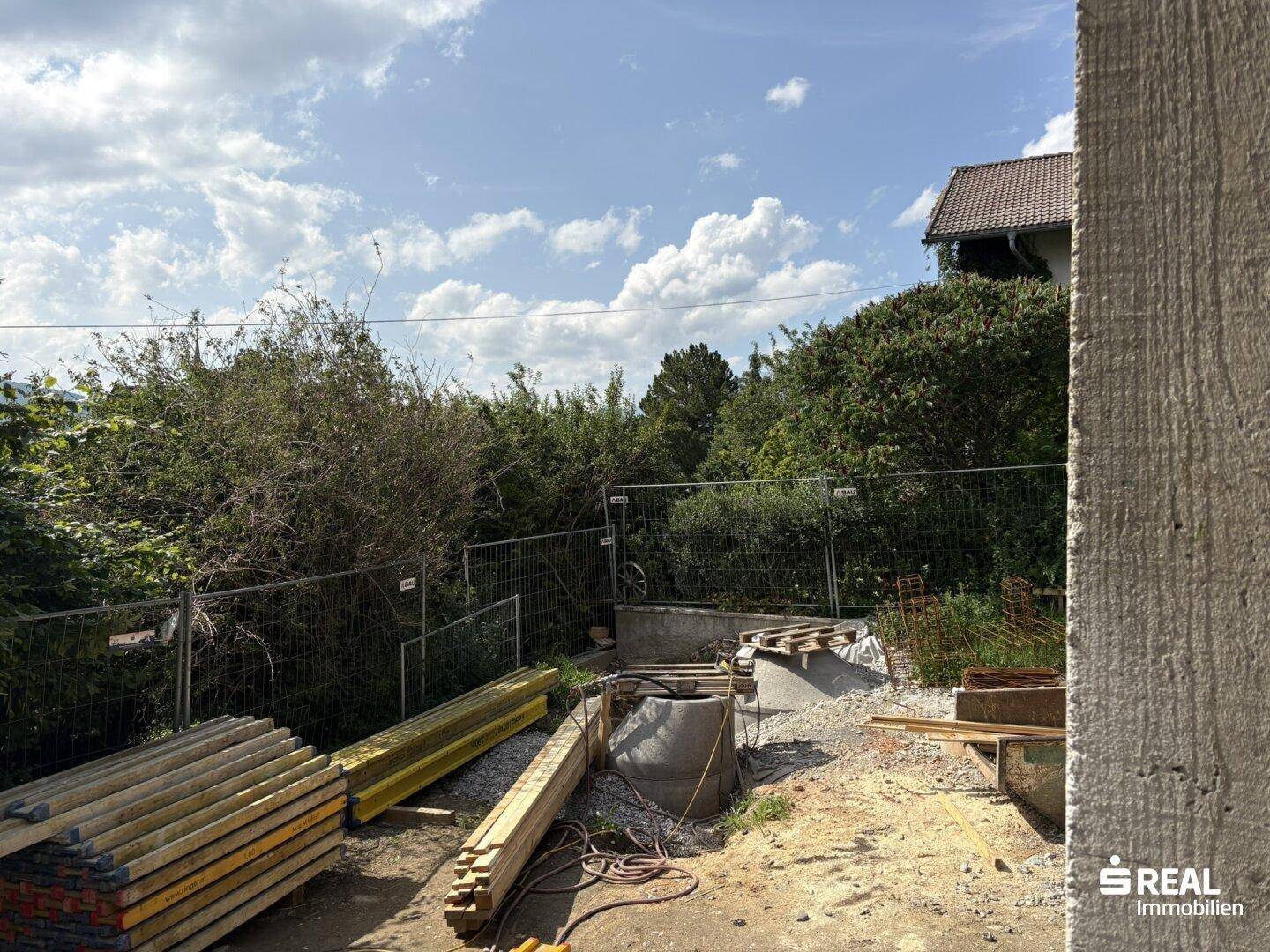 Baustelle mit verschiedenen Baumaterialien, einem Bauzaun und dichtem Buschwerk im Hintergrund unter blauem Himmel.