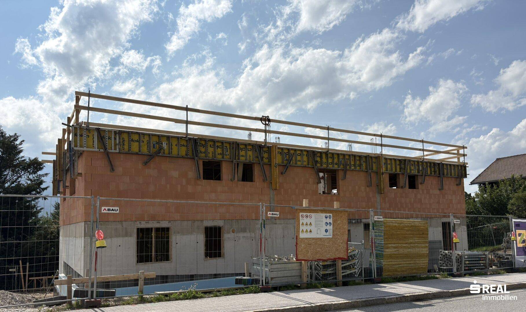 Neubau im Rohbauzustand mit sichtbaren Ziegelwänden und Betonelementen, umgeben von Baugerüst und Baustellenschildern.