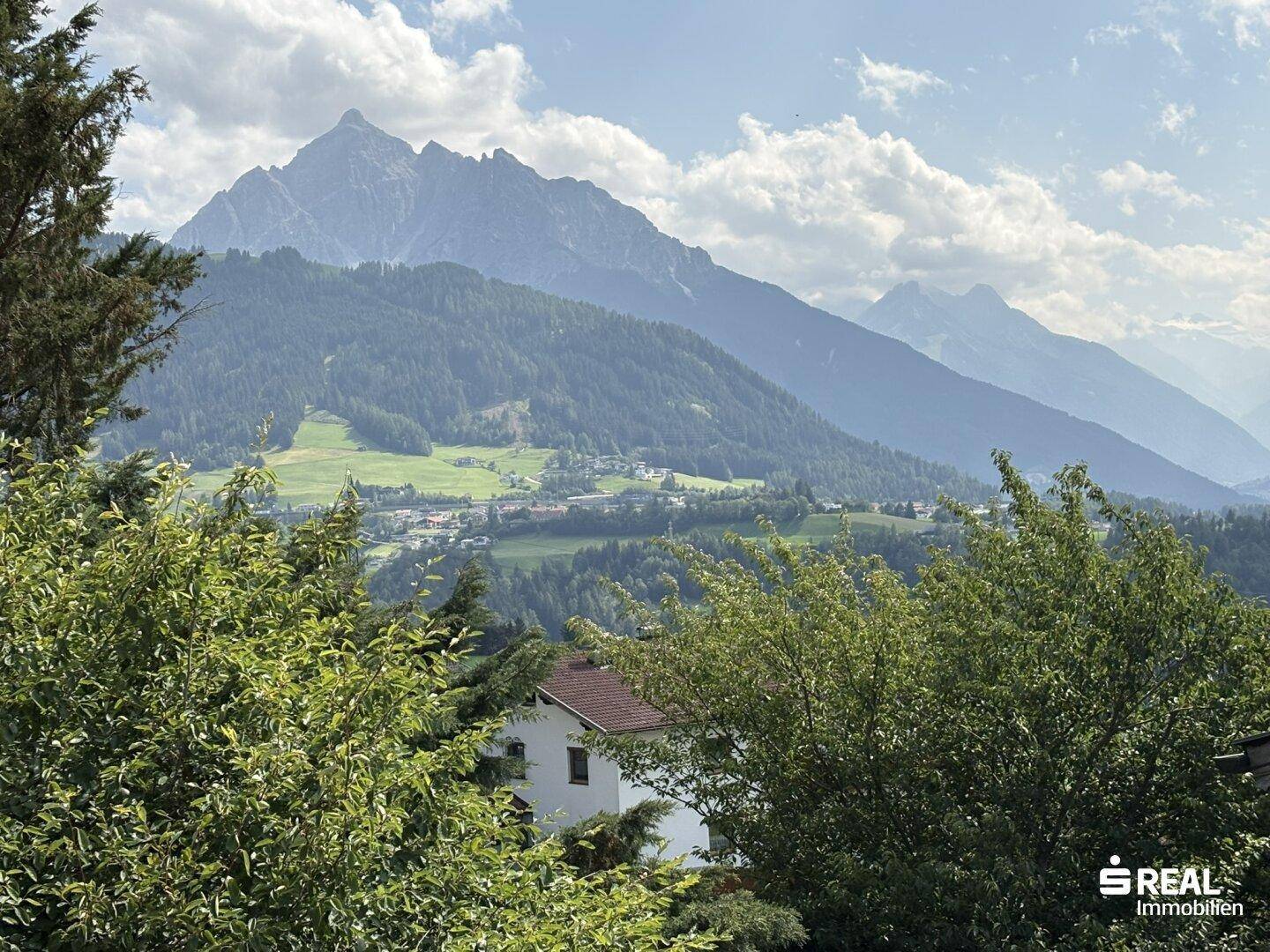 Panoramablick auf eine weite Berglandschaft mit grünen Tälern, Wäldern und vereinzelten Häusern unter bewölktem Himmel.
