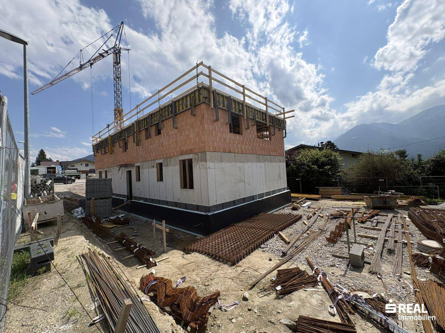 Neubau im Rohbau mit Fundament und Wänden, umgeben von Baumaterialien und einem Kran, mit Bergen im Hintergrund.