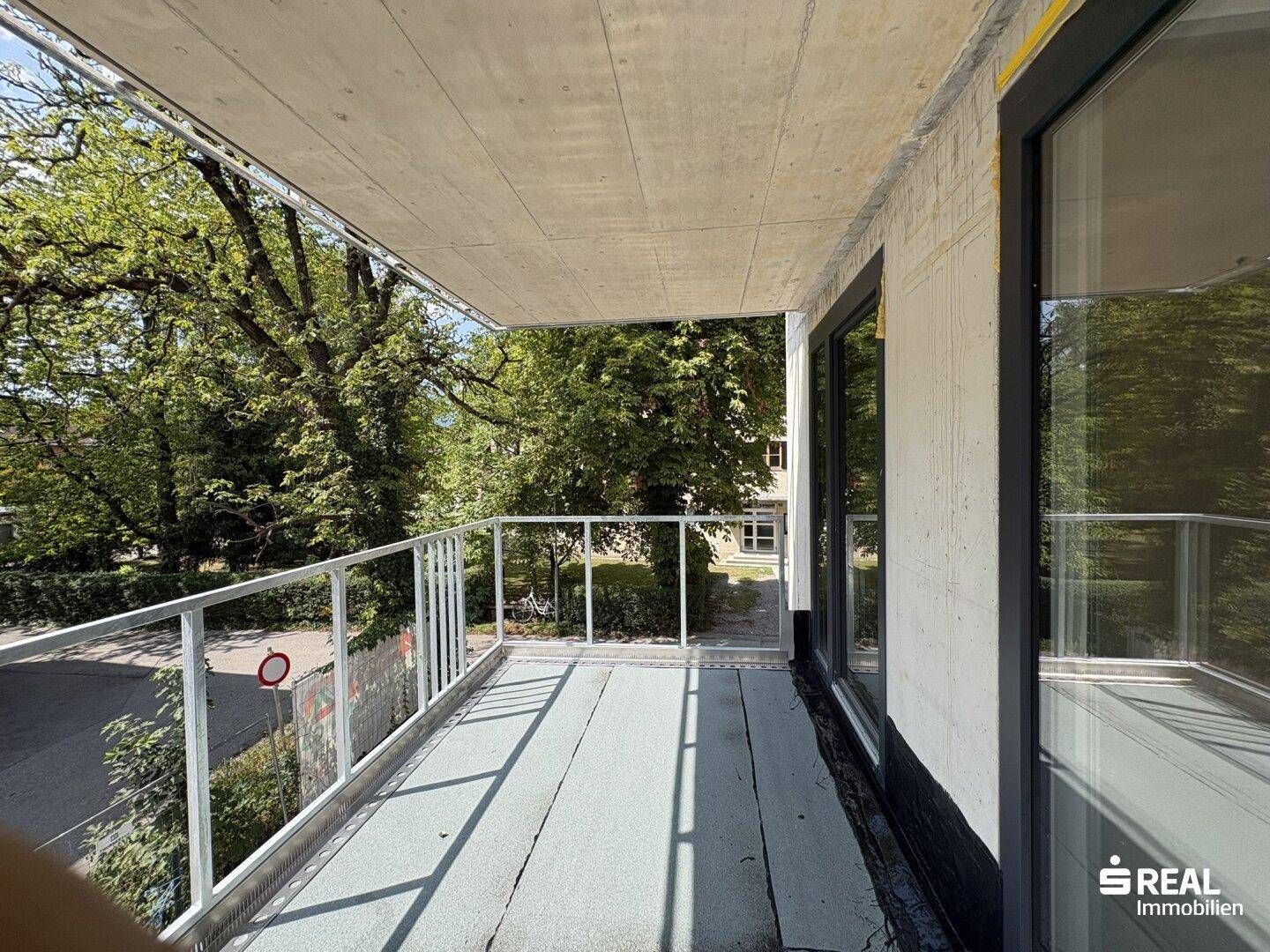 Unfertiger Balkon eines Neubaus mit Blick auf grüne Bäume und viel Tageslicht.