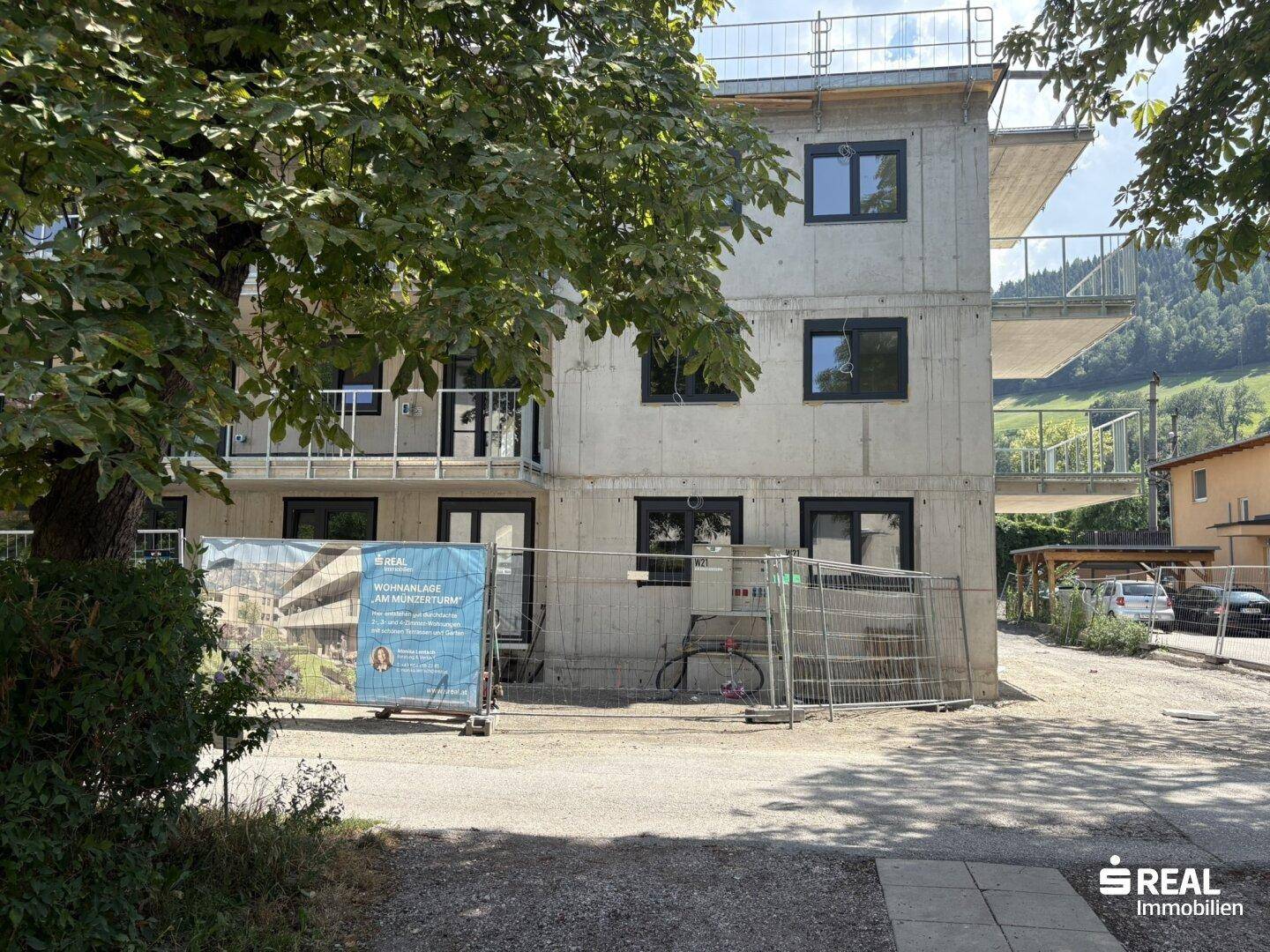 Neubau in Rohbauweise mit Betonfassade und Bauzaun, umgeben von Bäumen und Hügeln.