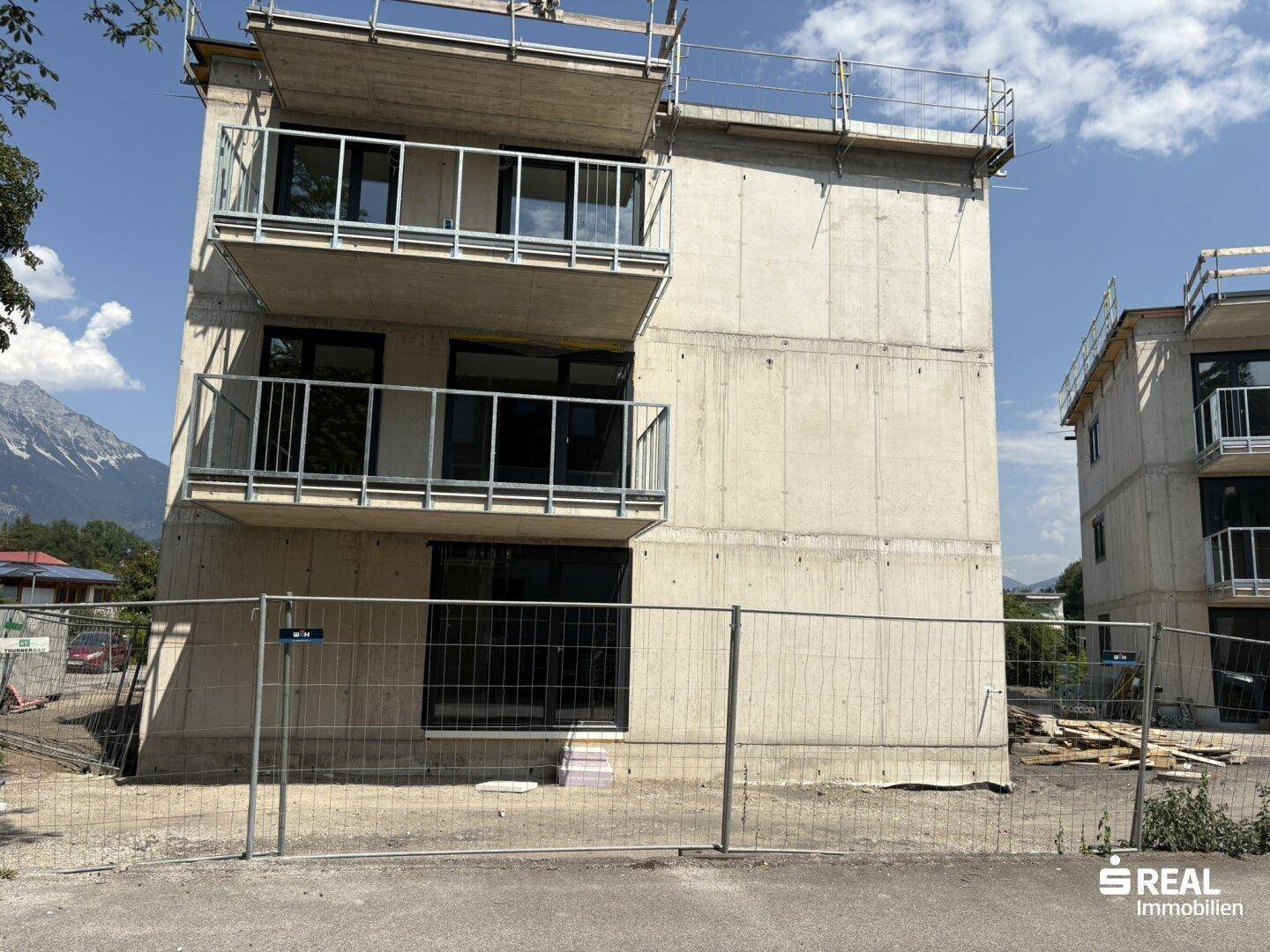 Neubau in Rohbauweise mit Betonfassade und Balkonen, umgeben von Bauzäunen und Bergen.