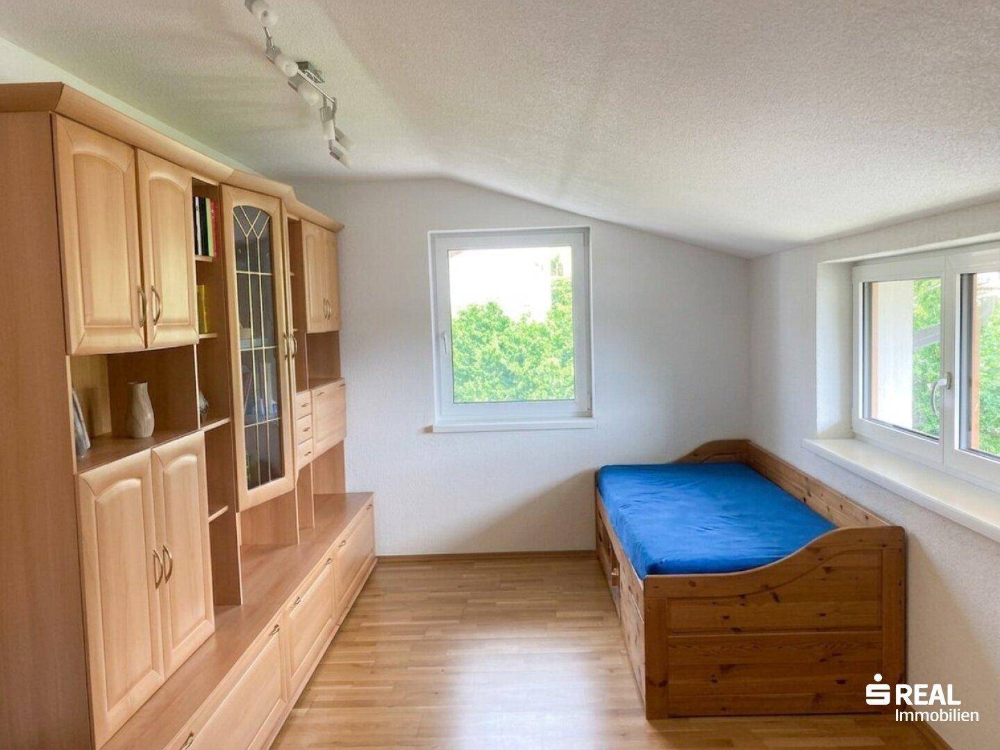 Helles Schlafzimmer mit Holzmöbeln, Laminatboden und Fenstern mit Blick ins Grüne.
