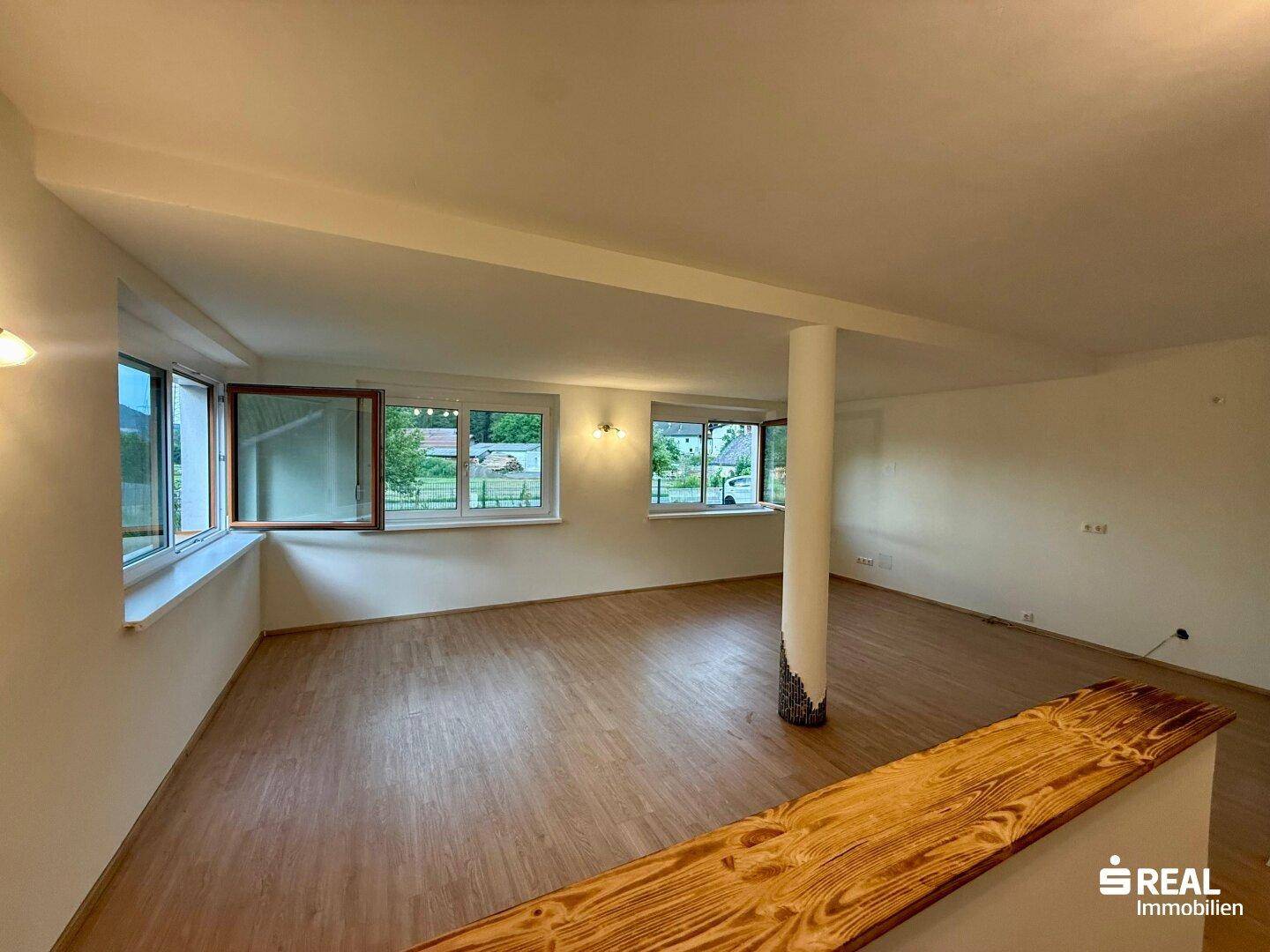 Geräumiger Wohnbereich mit Laminatboden und großen Fenstern, die viel Licht hereinlassen.