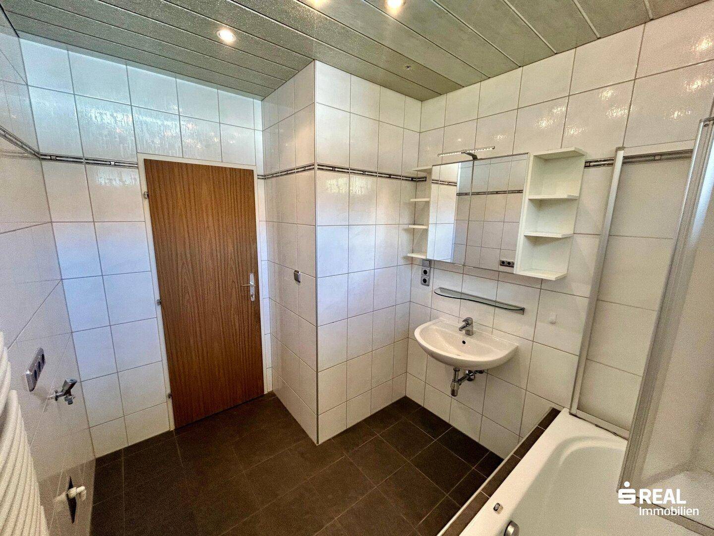 Badezimmer mit weißen Fliesen, Waschbecken, Badewanne mit Duschabtrennung und Spiegelschrank.