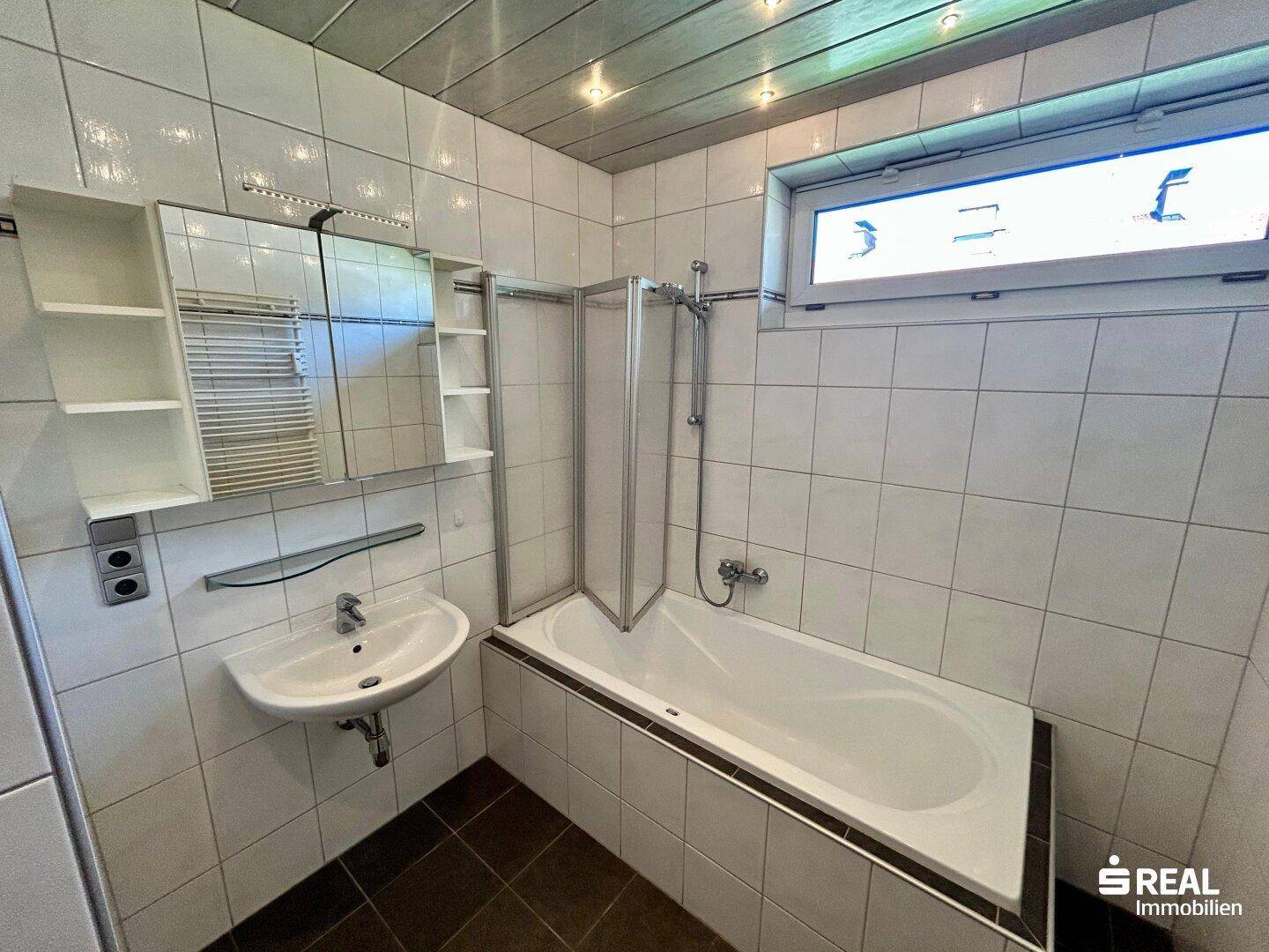 Funktionales Badezimmer mit Badewanne, Duschabtrennung, Waschbecken und Fenster für natürliche Belüftung.