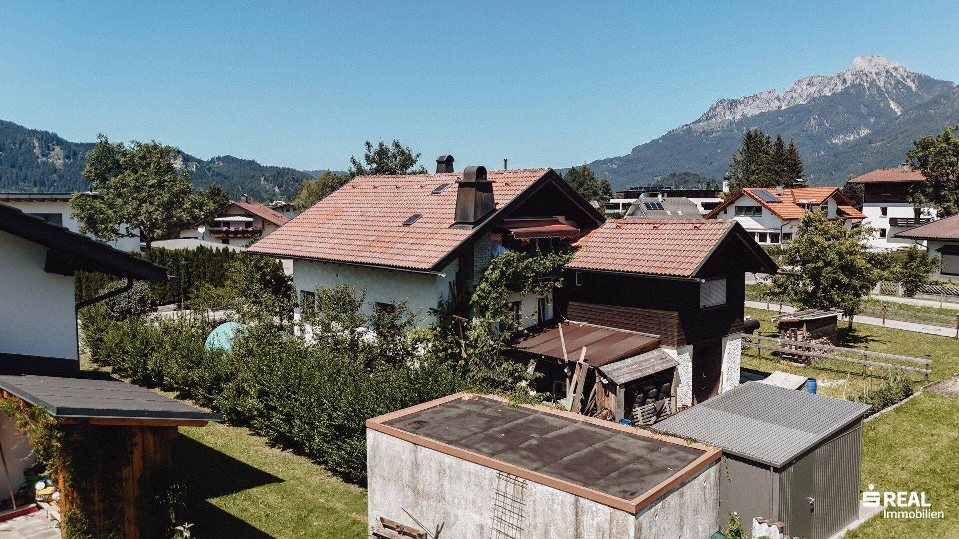 Seitenansicht des Hauses mit gepflegter Hecke, Nebengebäuden und einem weiten Blick auf die Berge.