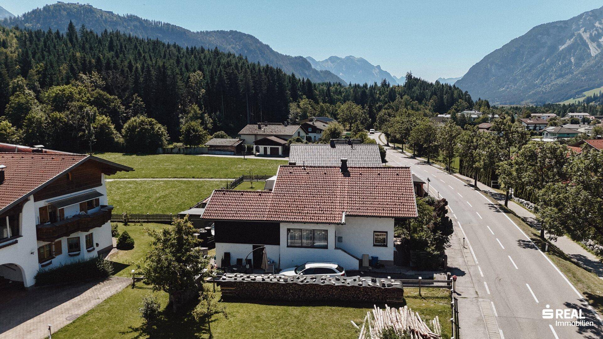 Weitläufige Luftaufnahme des Hauses mit angrenzender Straße und der beeindruckenden Bergkulisse.