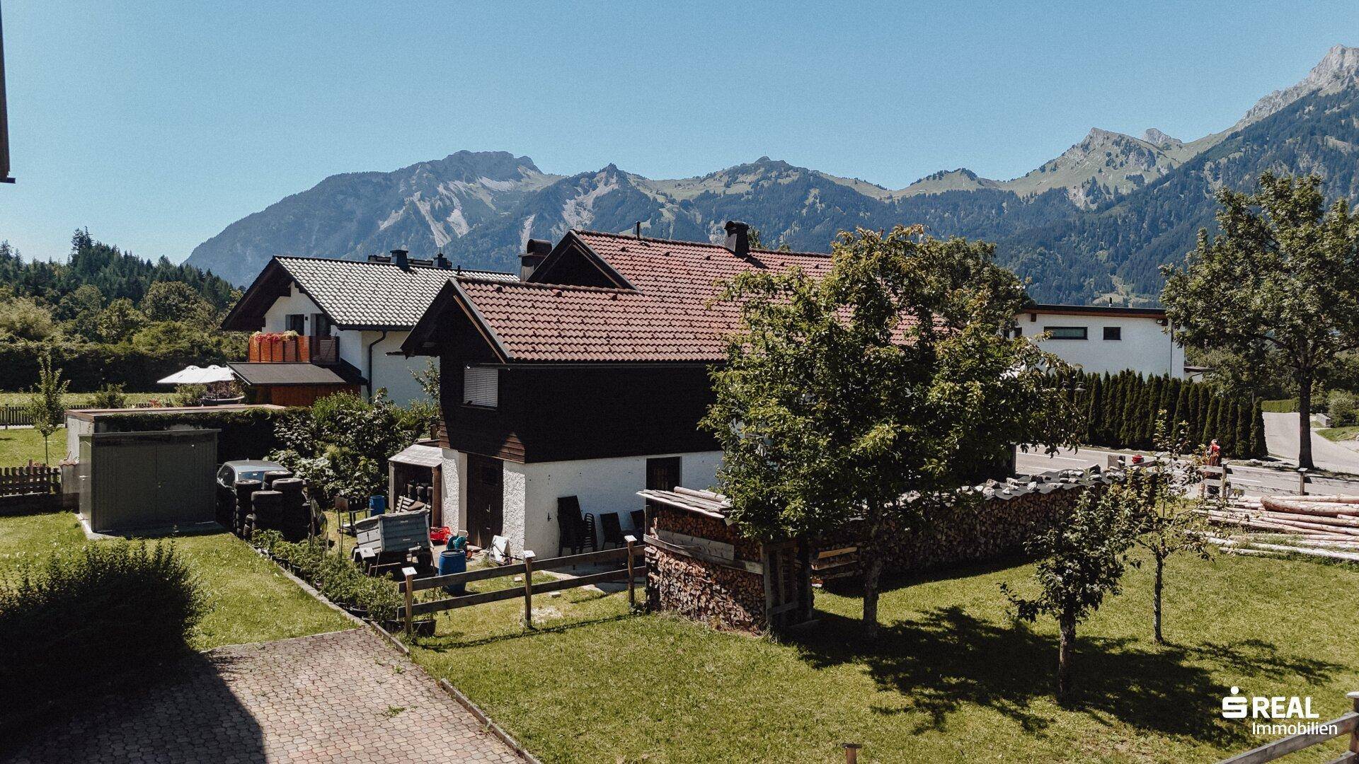 Sonniger Gartenbereich des Hauses mit Holzstapel und beeindruckendem Bergblick im Hintergrund.